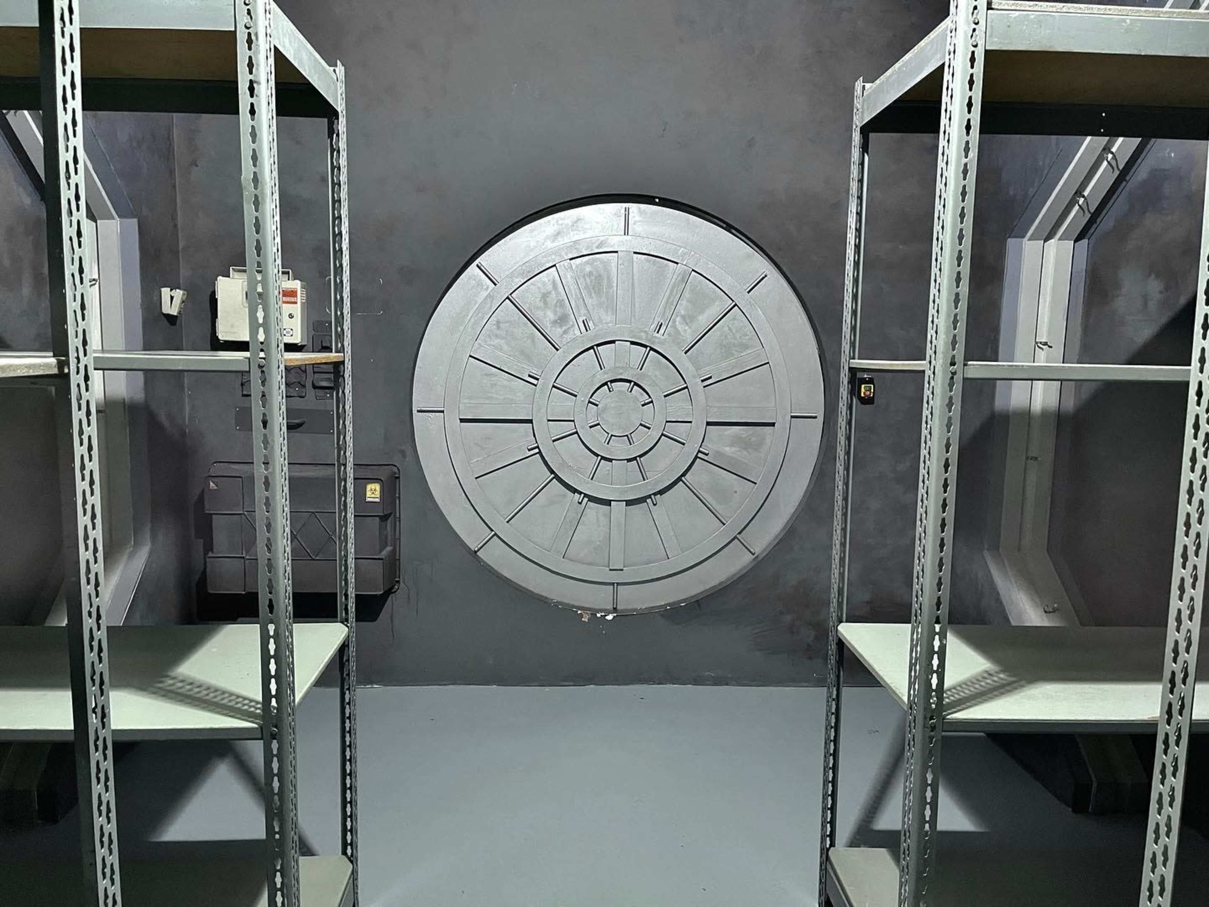 A dark room with metal shelving units on either side and a circular metallic door at the center of the back wall.