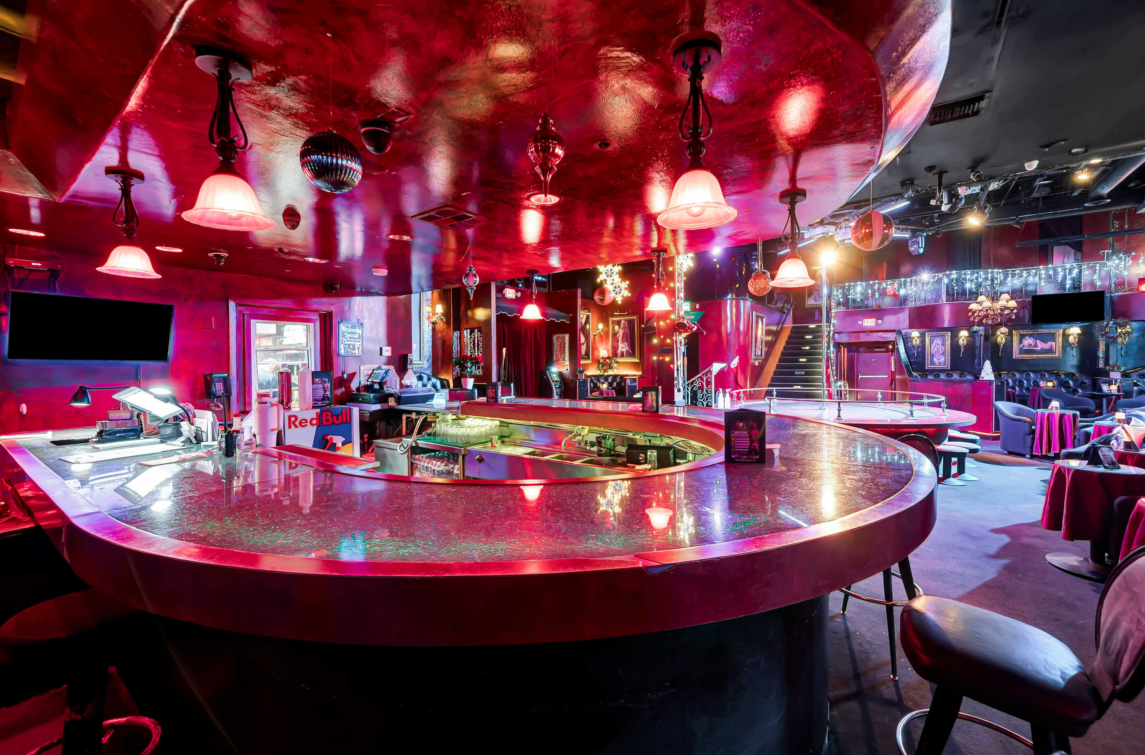 The image shows a dimly lit bar with a circular counter, red lighting, and eclectic decor, including chandeliers and framed art.