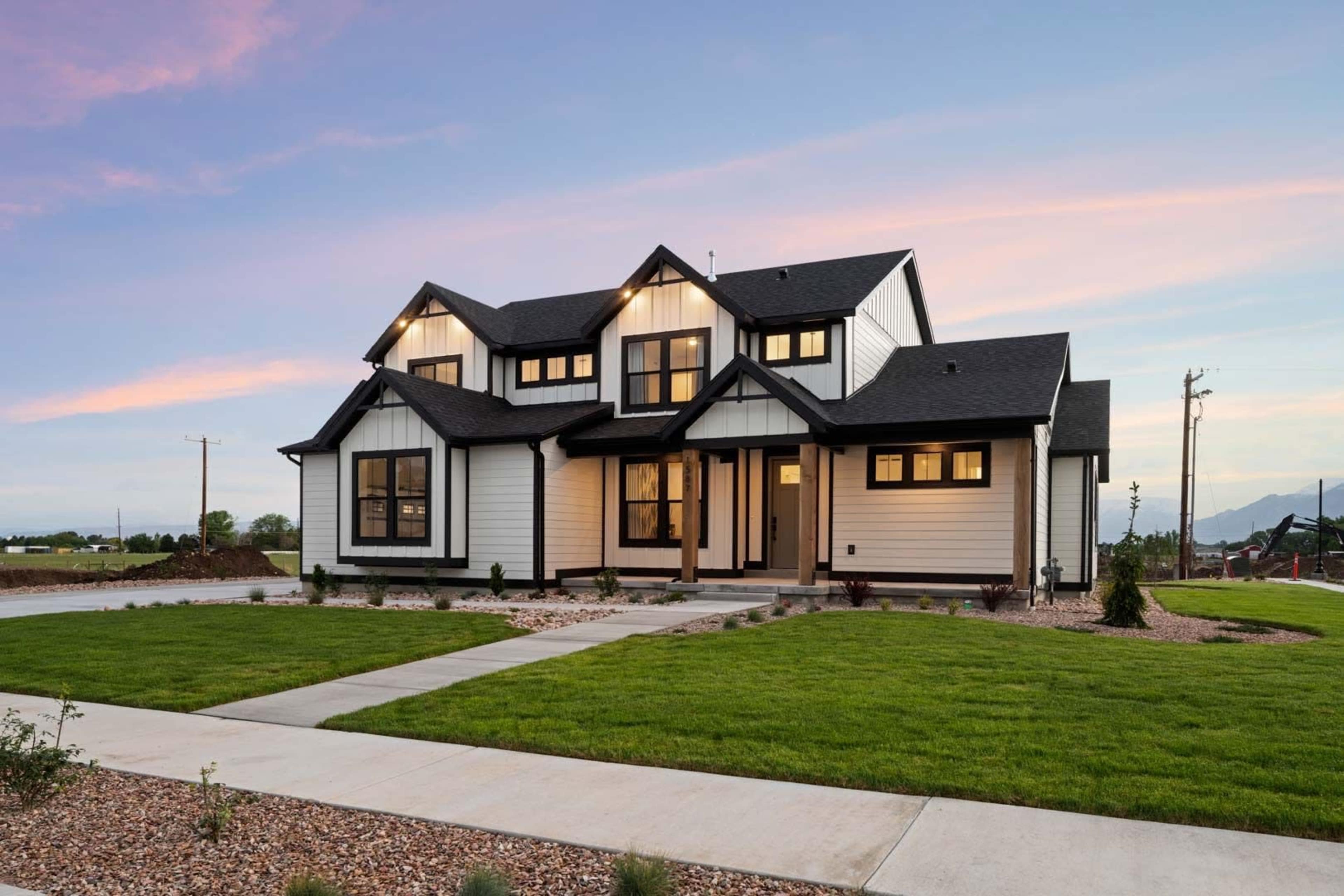 A large, modern house with a white exterior and black accents sits on a landscaped lot, featuring a wide porch and multiple windows, against a backdrop of a clear sky.