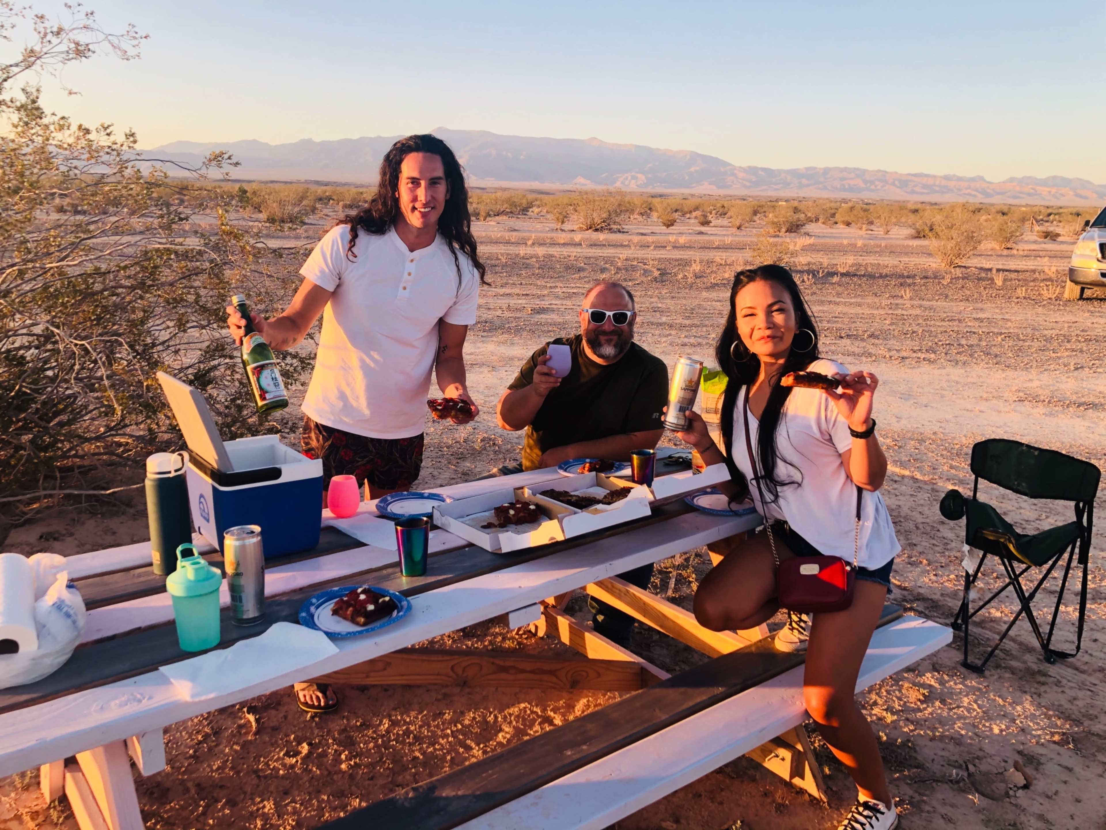 Three people hold drinks and snacks while posing at a picnic table in a desert landscape during sunset.