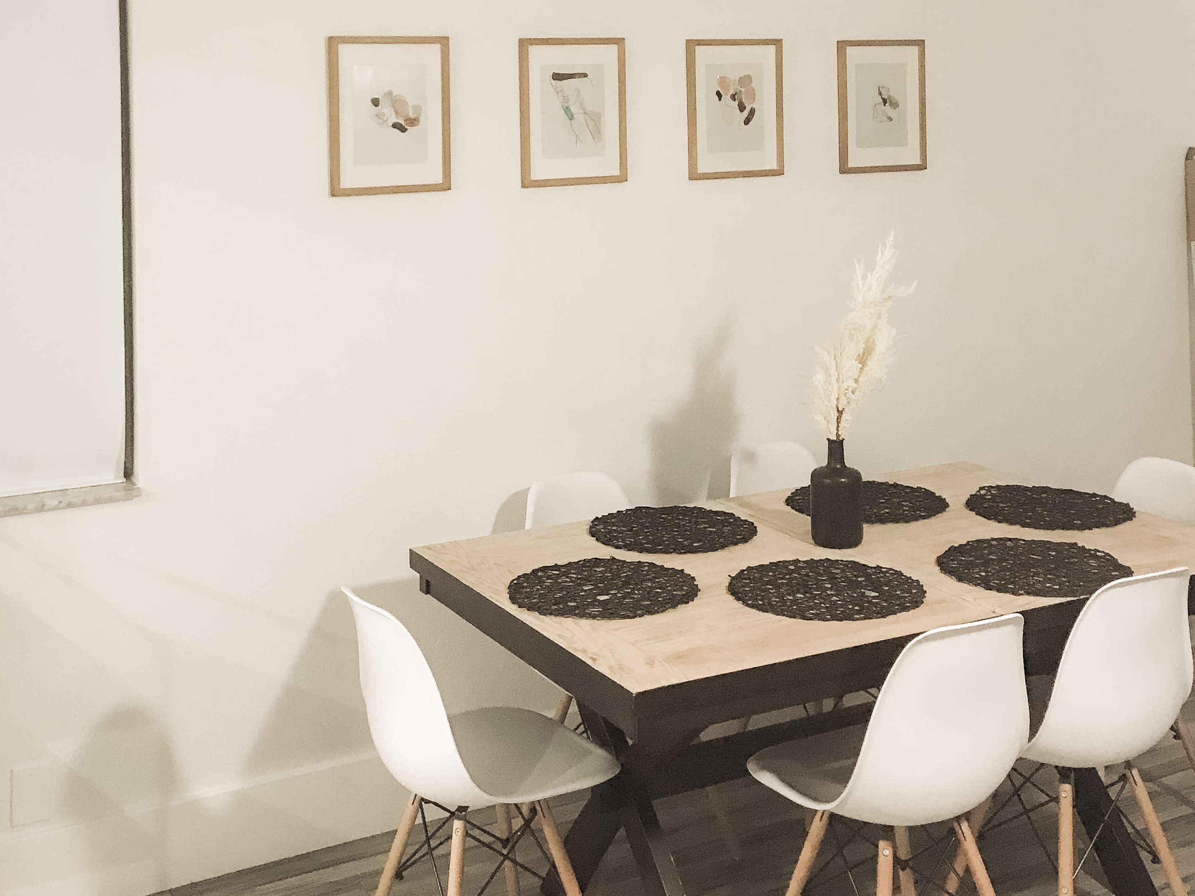 A dining table with a wooden top, surrounded by white chairs, is set with black placemats and a vase containing dried flowers, against a wall adorned with framed artwork.
