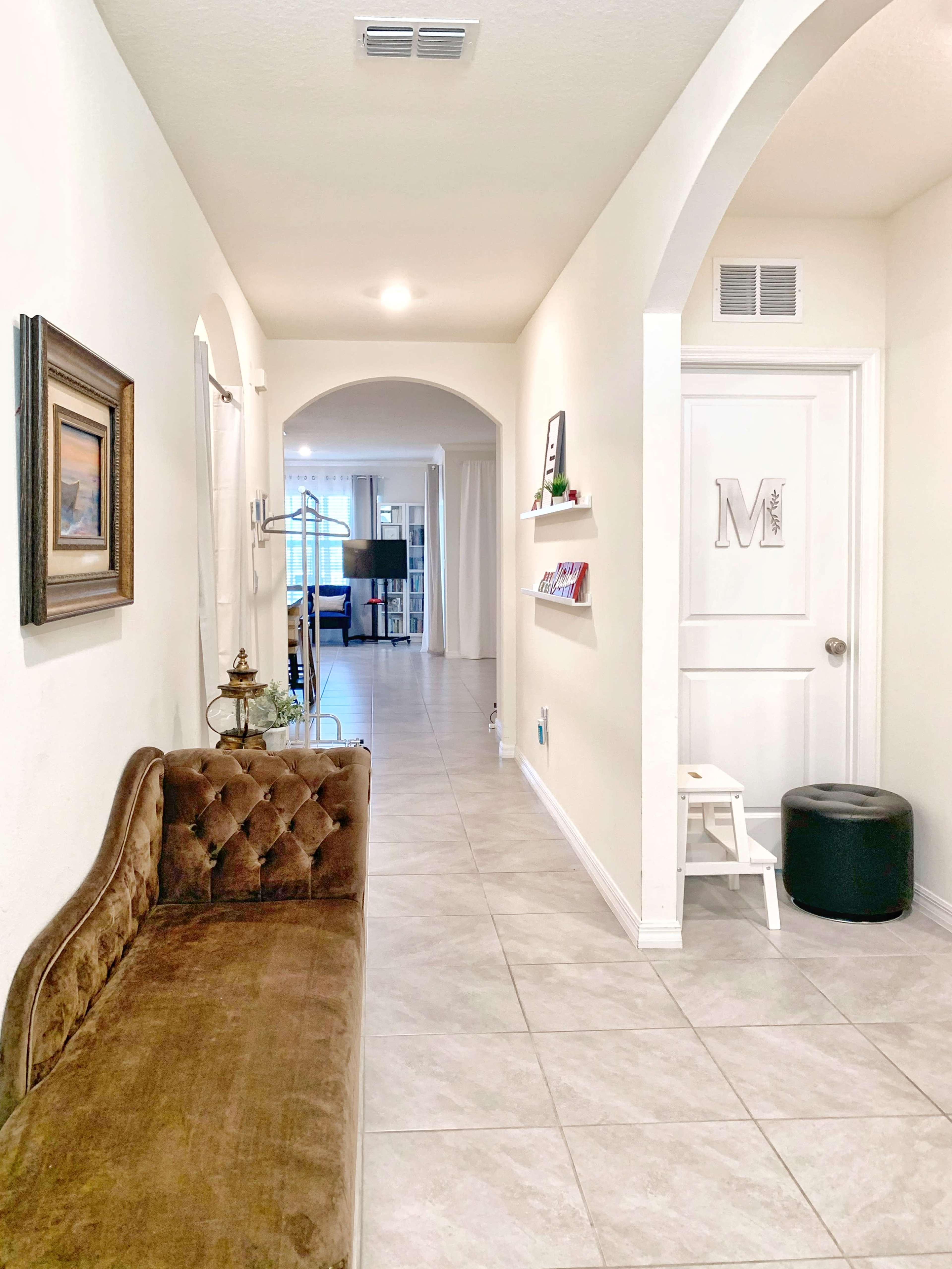 The image shows a corridor with a brown tufted bench on one side, leading to an arched doorway at the end and a closed door with a monogram.