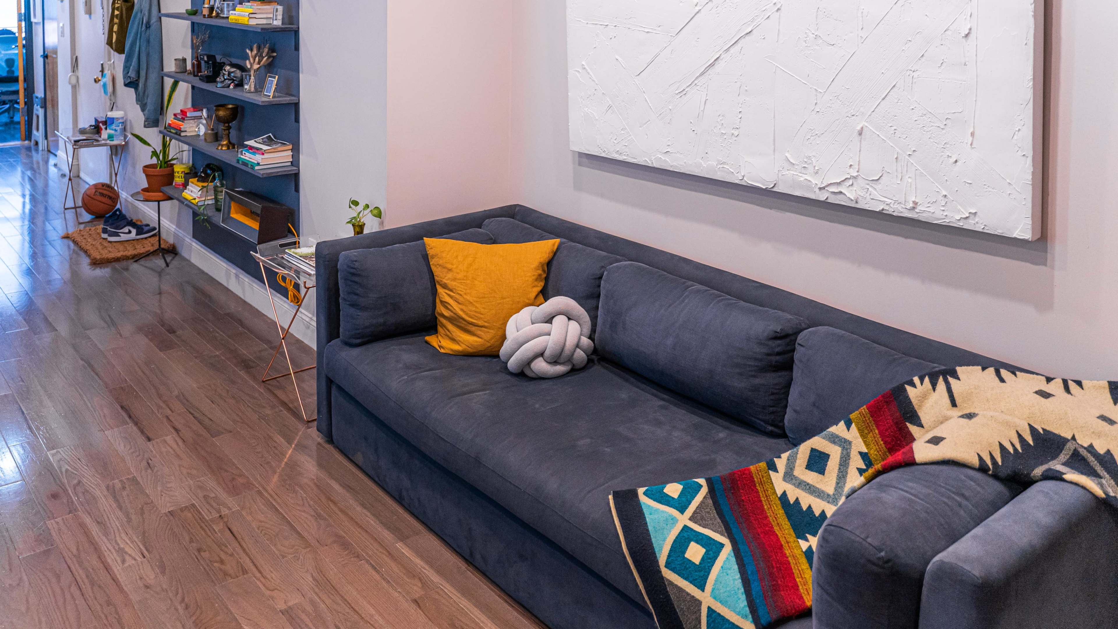 A blue sofa with a yellow cushion and a decorative knot pillow sits against a light-colored wall, accompanied by a patterned throw blanket and a bookshelf filled with objects.