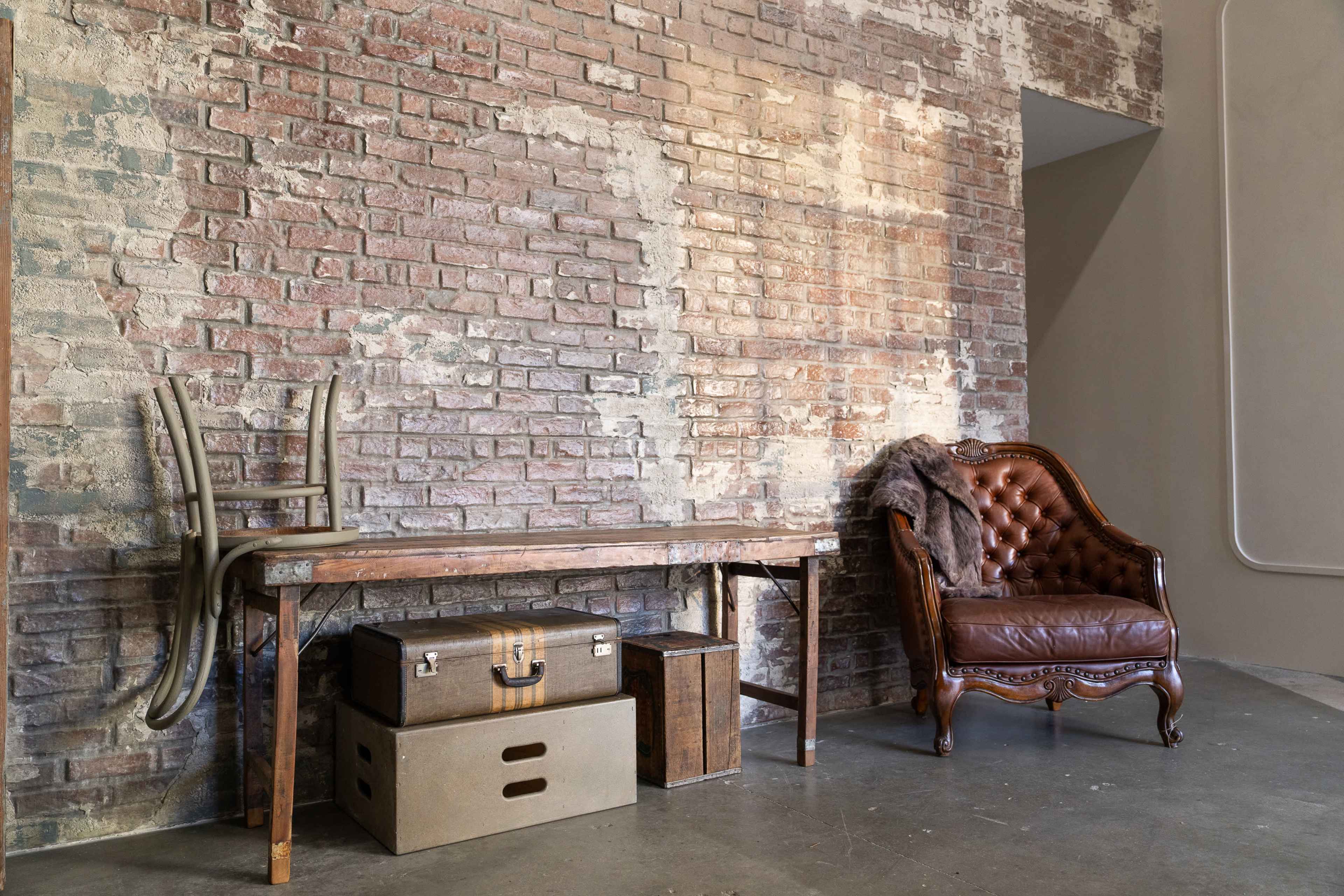 A wooden table and a leather armchair are positioned against a weathered brick wall, with a stack of storage boxes on the floor.