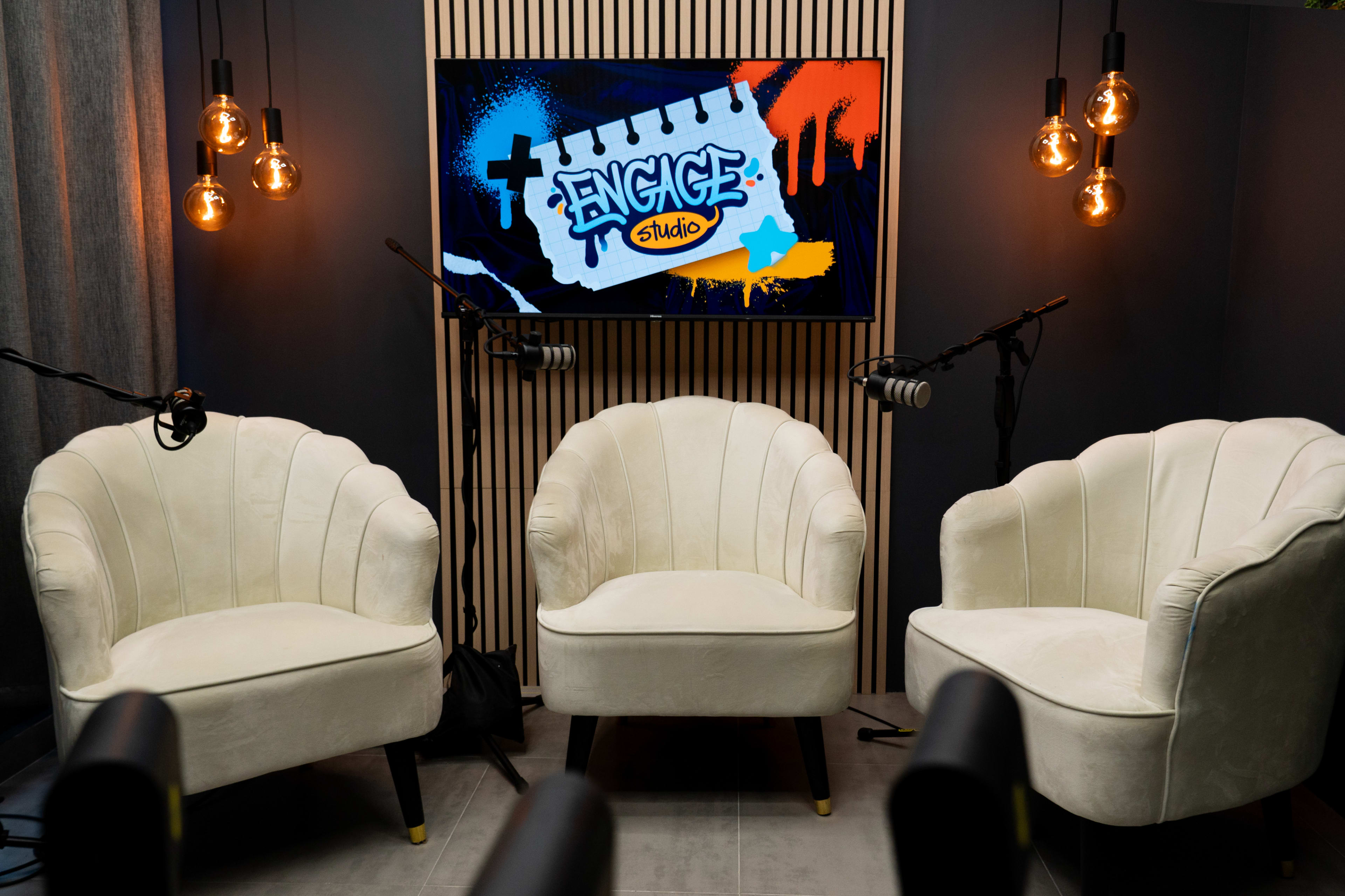The image shows a modern studio setup with three white chairs, a large screen displaying "ENGAGE Studio," and pendant lights hanging from the ceiling.