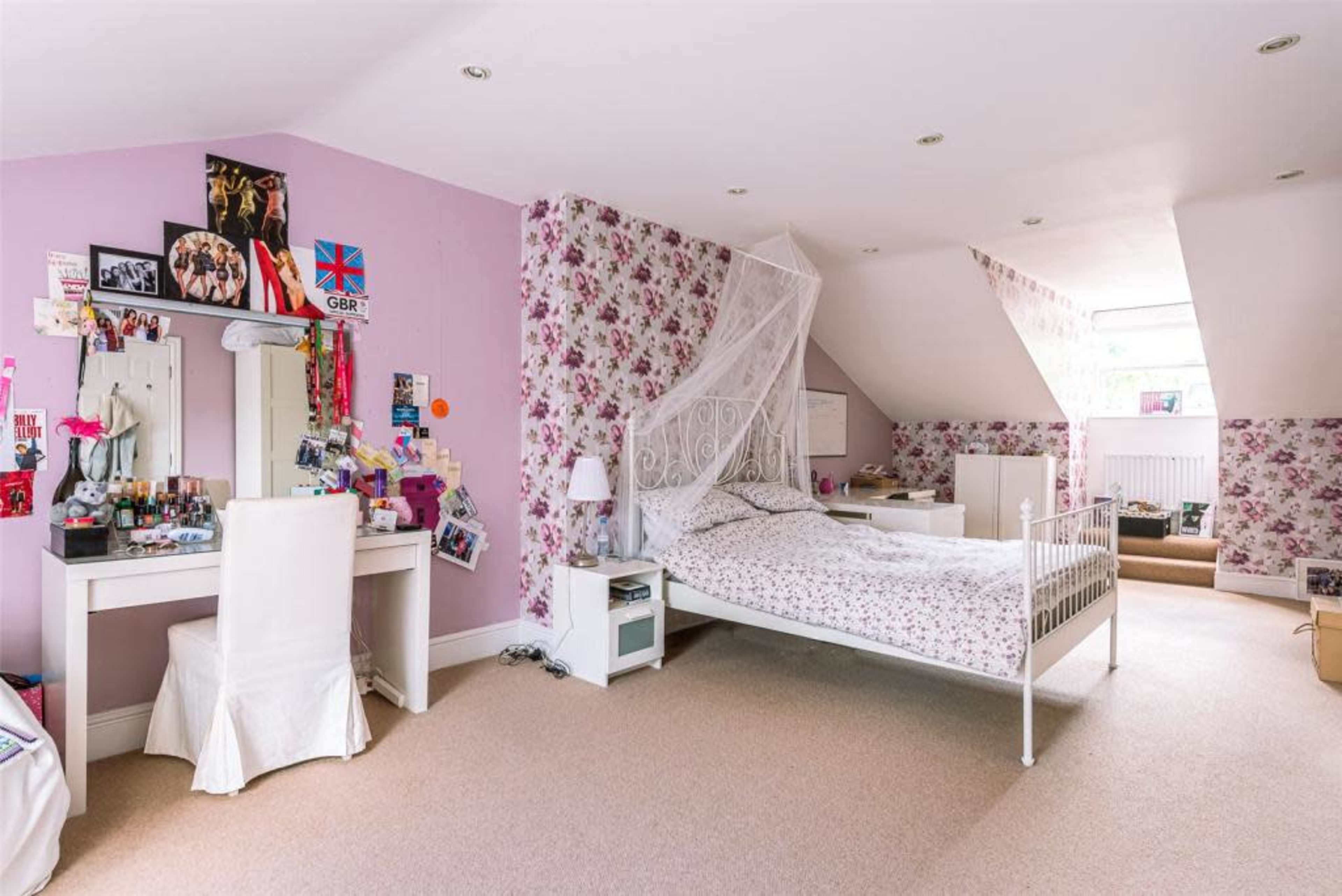 The image shows a bedroom with floral wallpaper, a bed with a patterned duvet, a vanity table with various cosmetics, and a large window providing natural light.