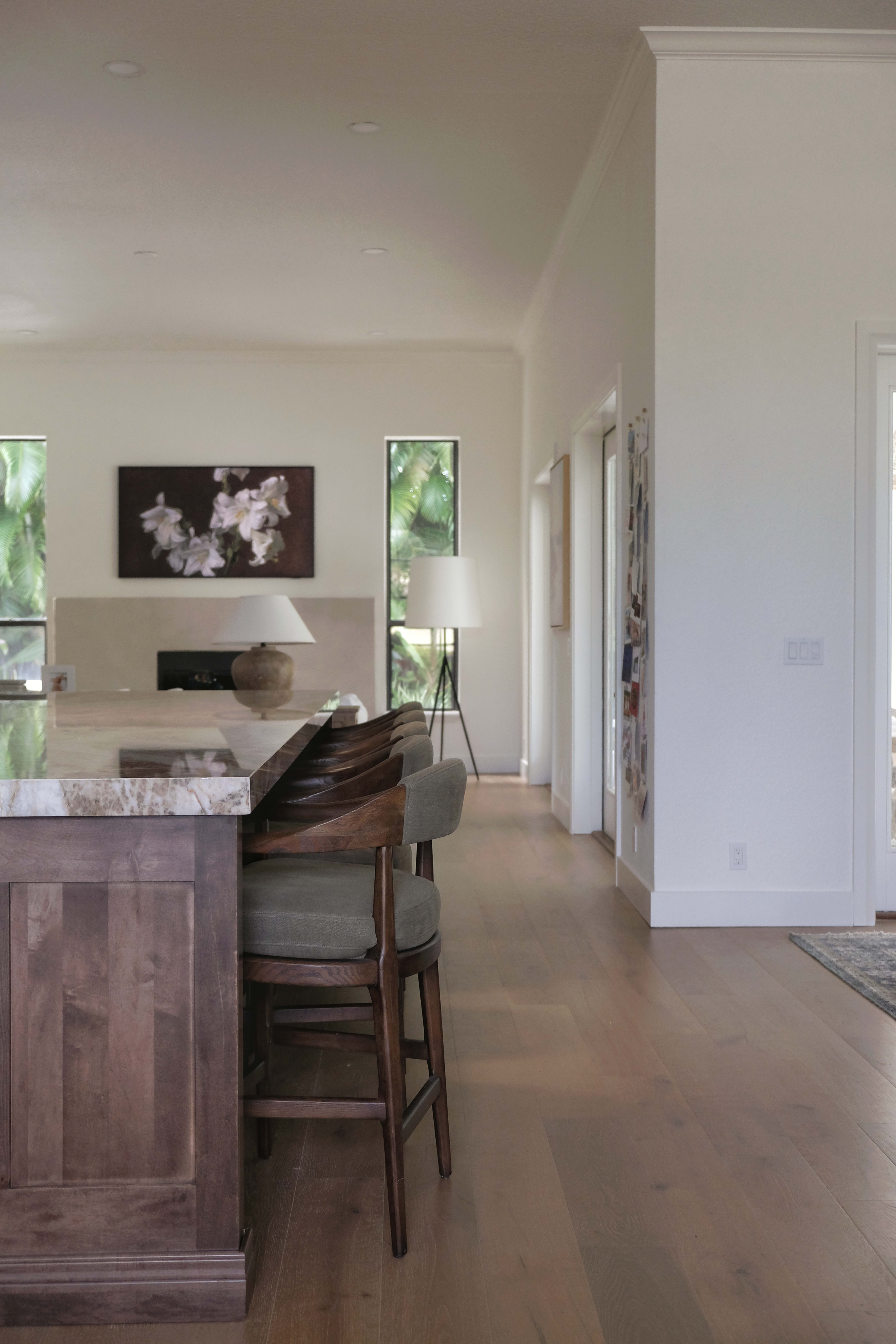 The image shows a modern kitchen featuring a wooden island with bar stools, a light-filled living area with large windows, and minimalist decor.
