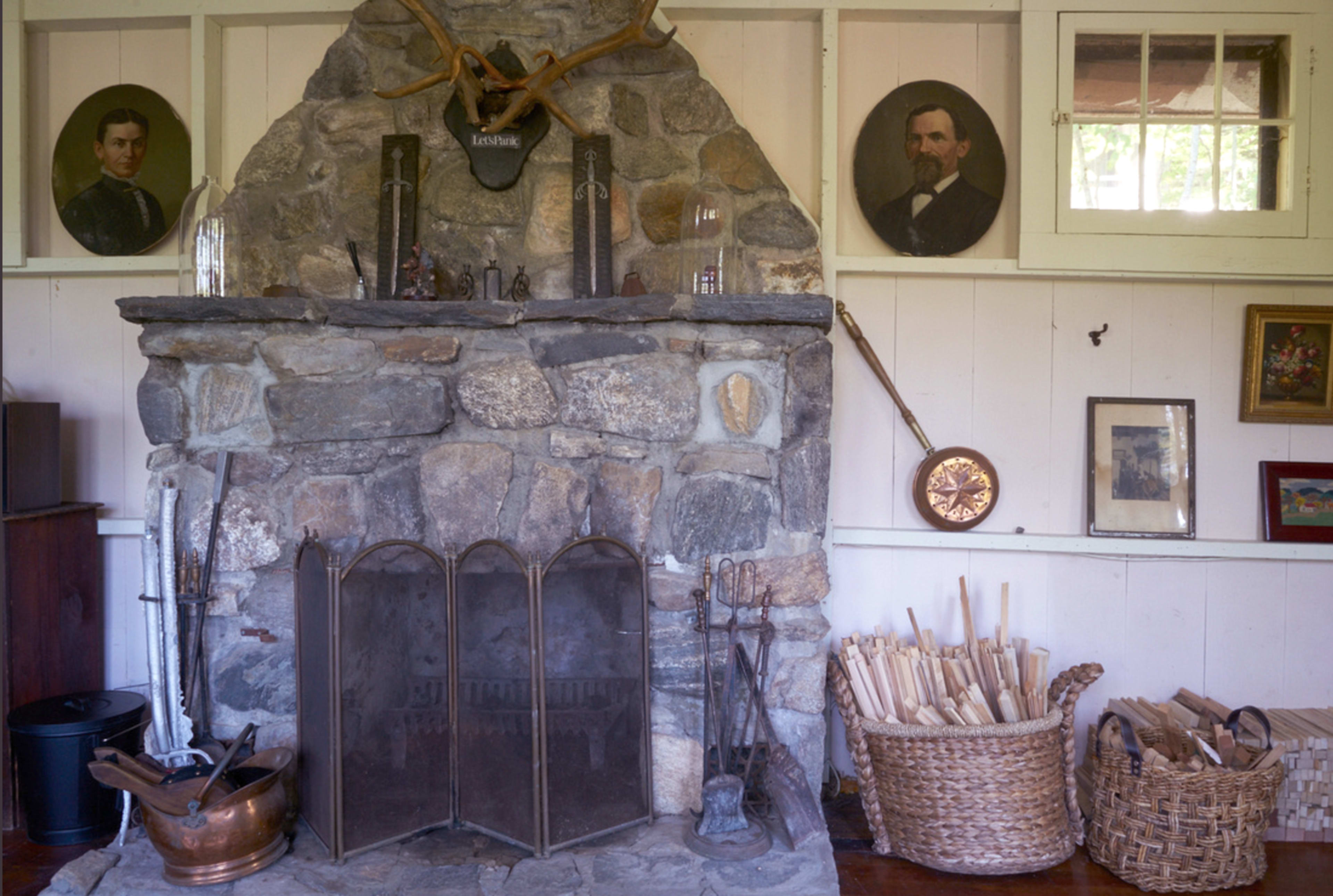 The image features a stone fireplace with decorative antlers above it, flanked by two portraits, and surrounded by various household items and a basket of wood.