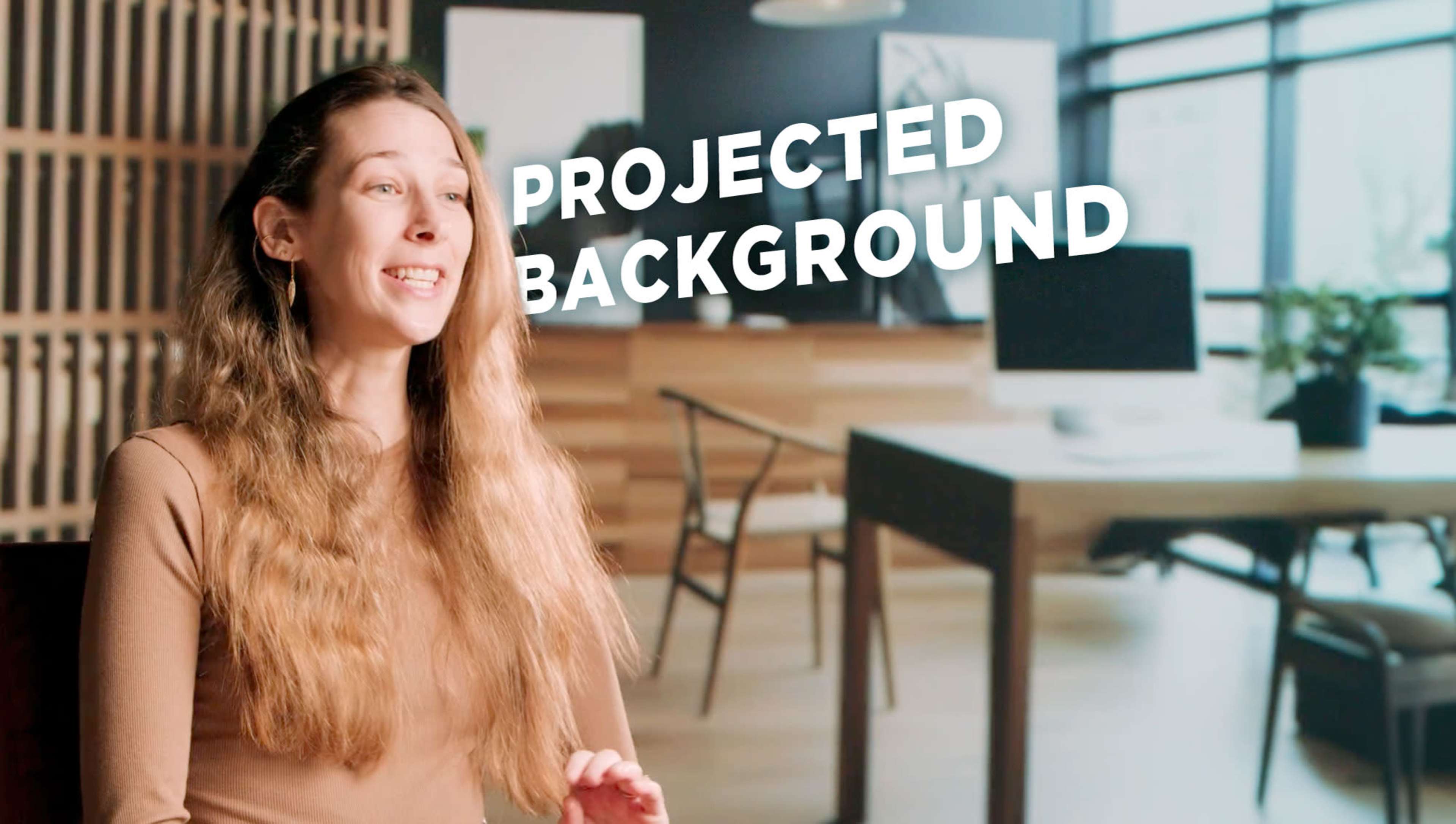 A woman with long hair sits in front of a modern office setting, with a projected background displaying the text "PROJECTED BACKGROUND."