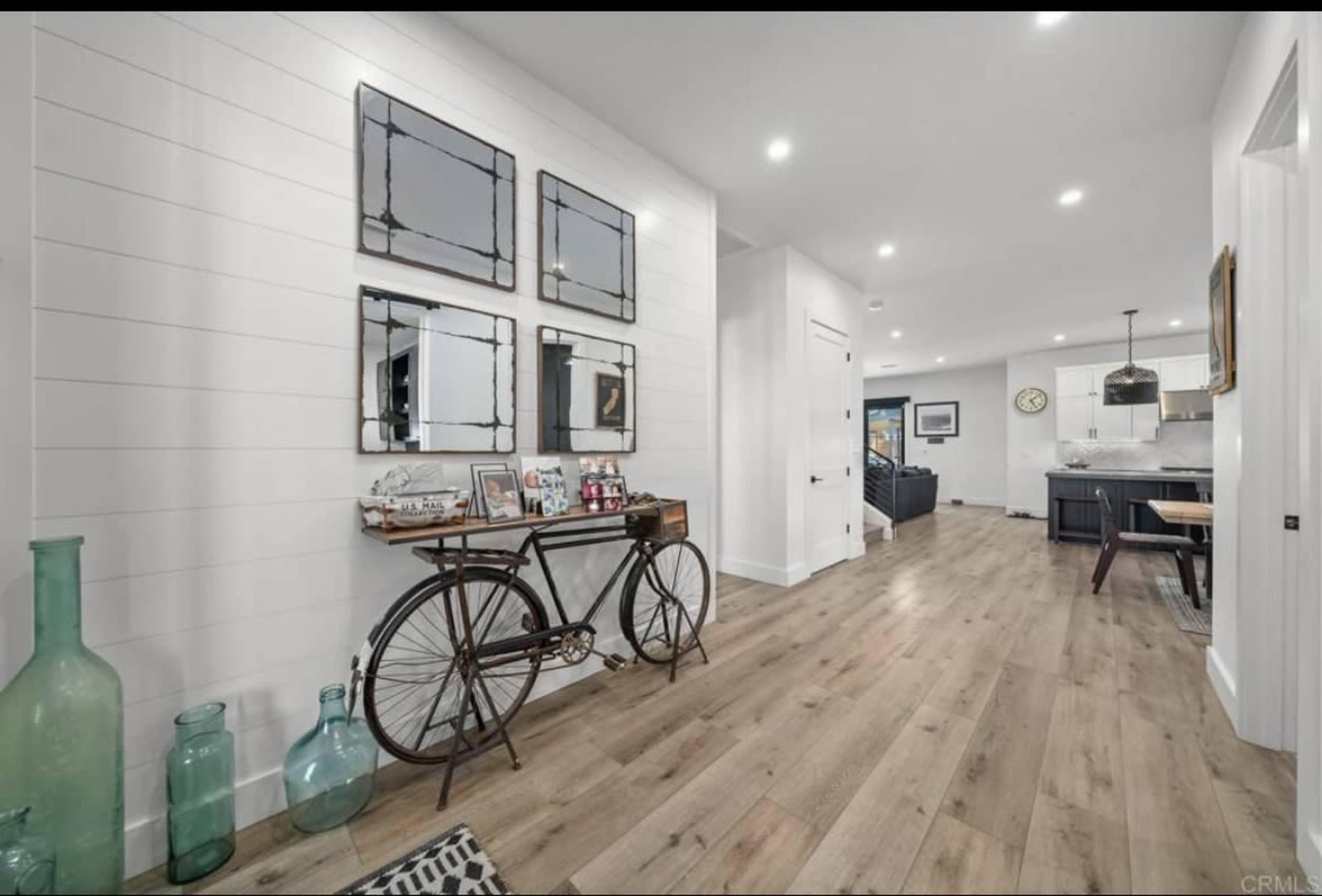 The image shows a hallway with light wooden flooring, a vintage bicycle display, decorative mirrors on the wall, and a glimpse of a living area and kitchen in the background.