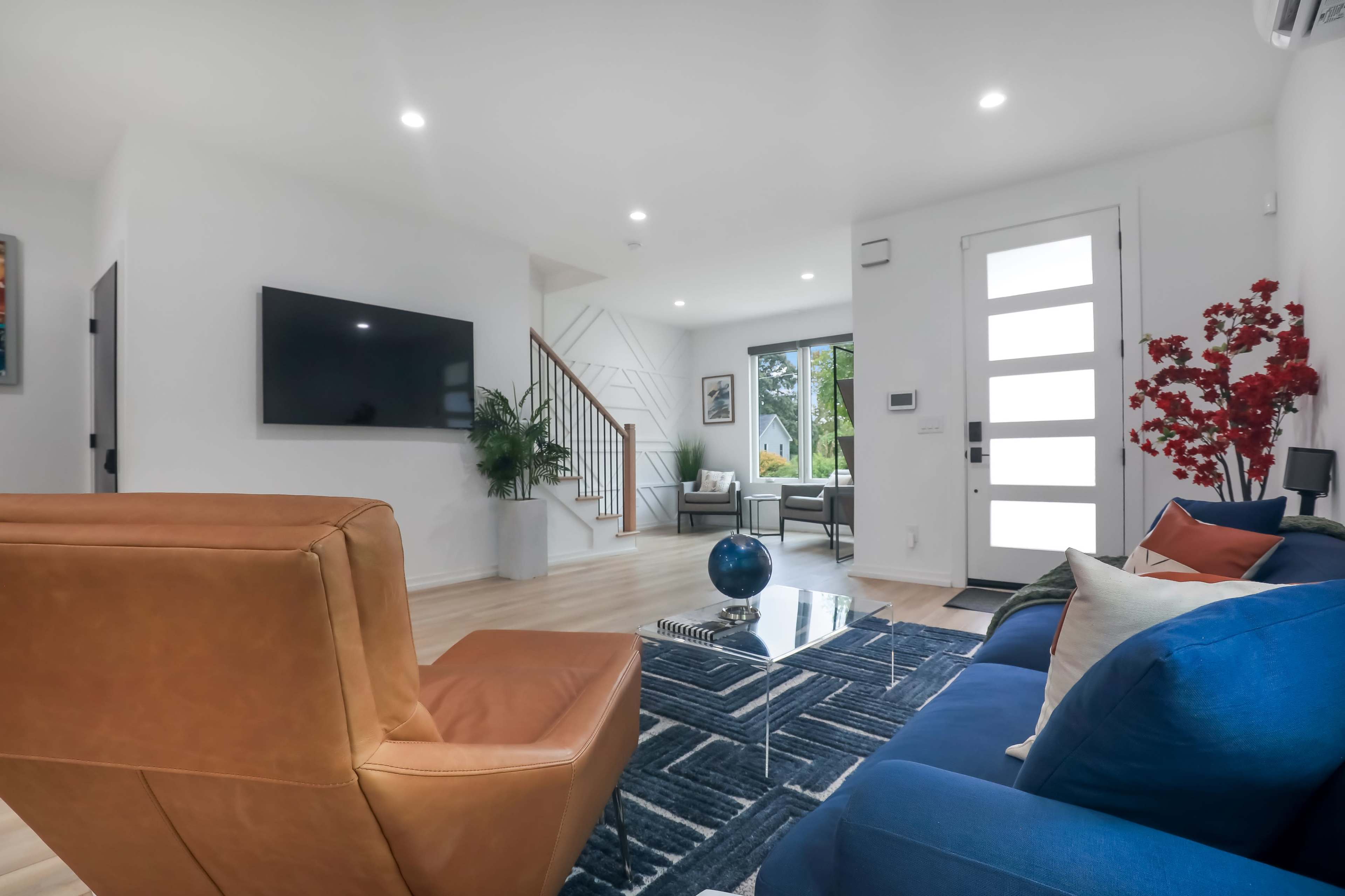 A modern living room with a large television mounted on the wall, a glass coffee table, and a blue and brown seating arrangement, leading to an entrance with a glass door.
