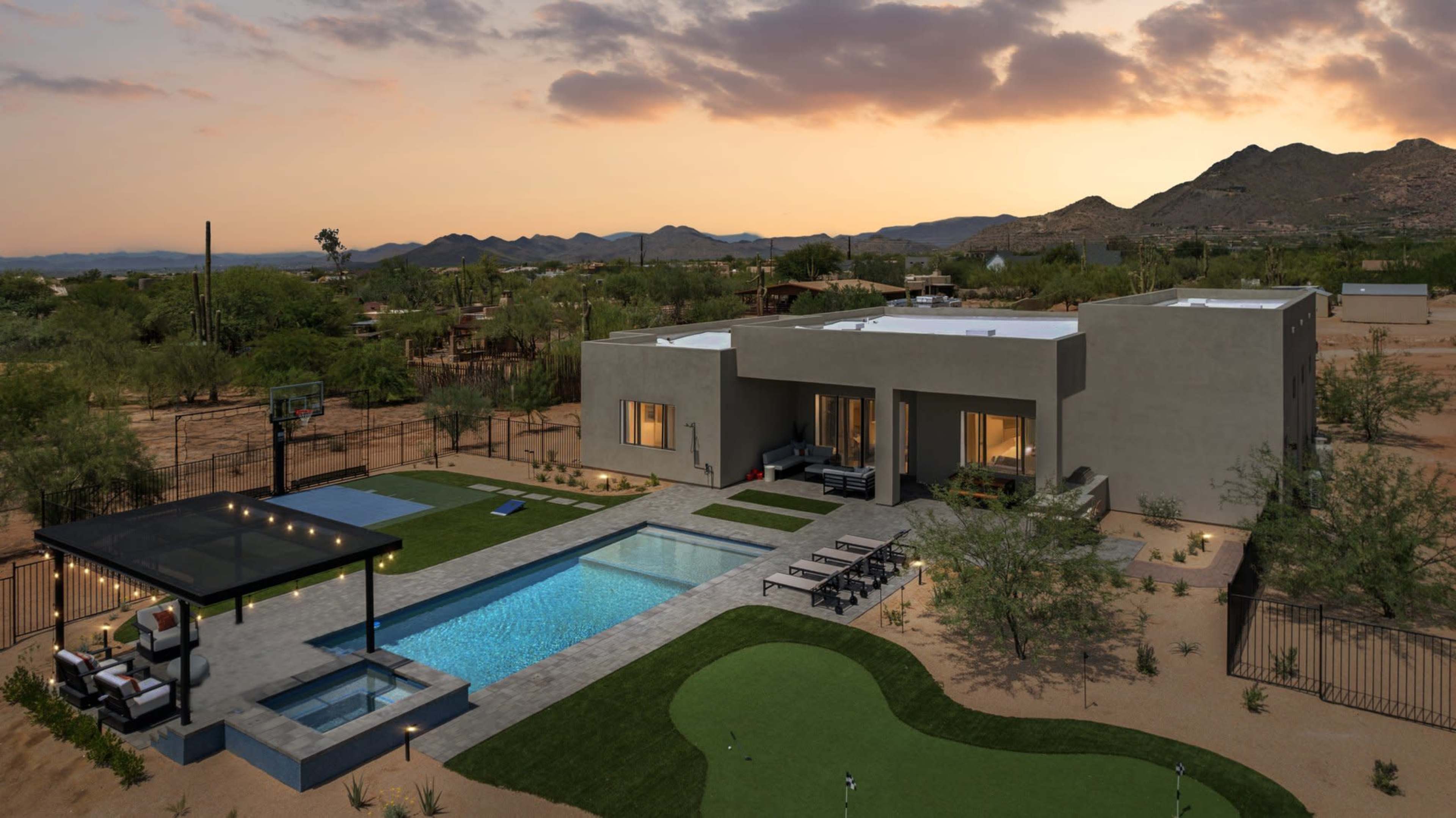 The image shows a modern single-story house with a swimming pool and a landscaped yard at sunset, surrounded by desert mountains.