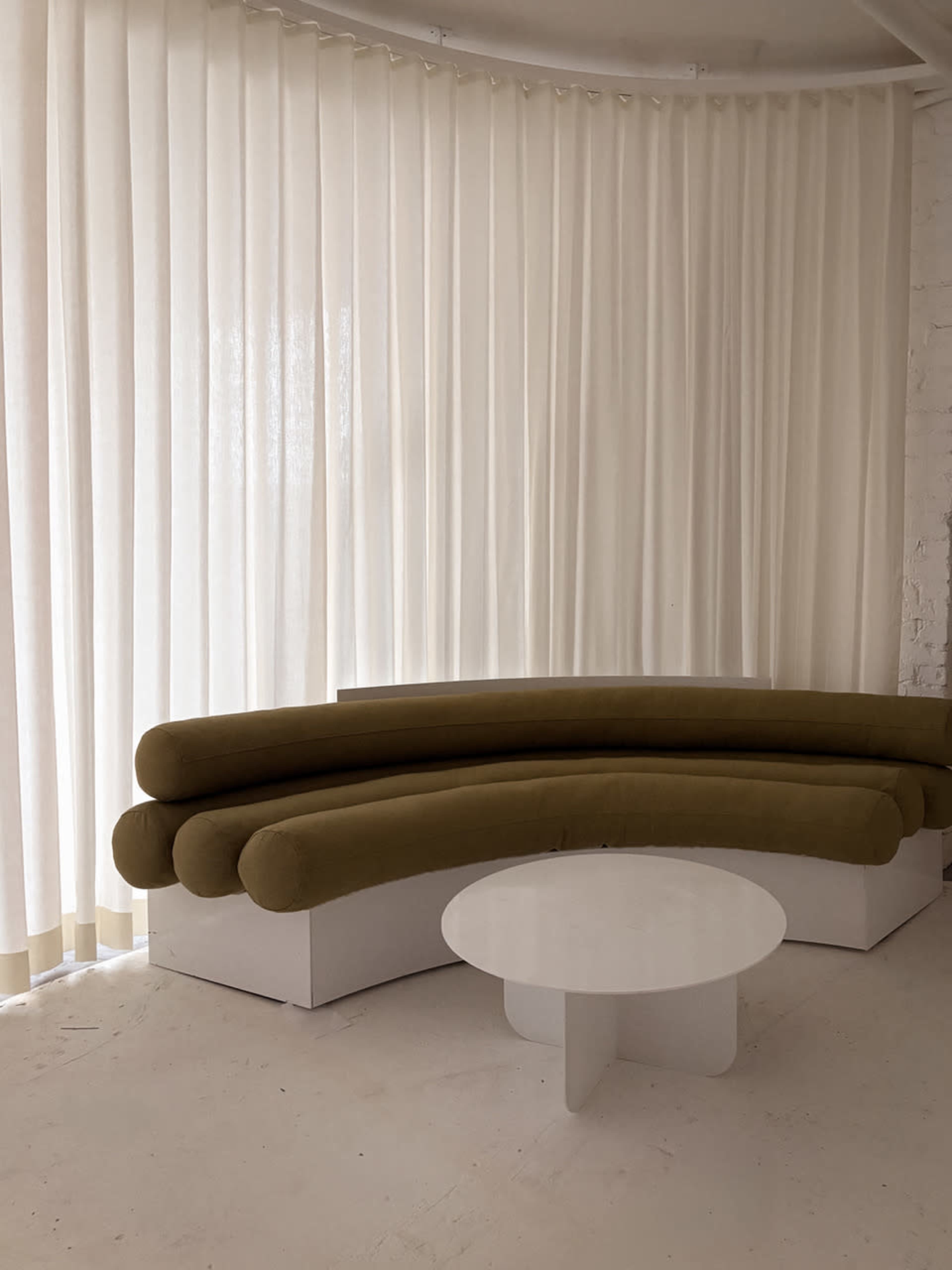 The image shows a curved green sofa against a backdrop of sheer white curtains, accompanied by a round white coffee table.