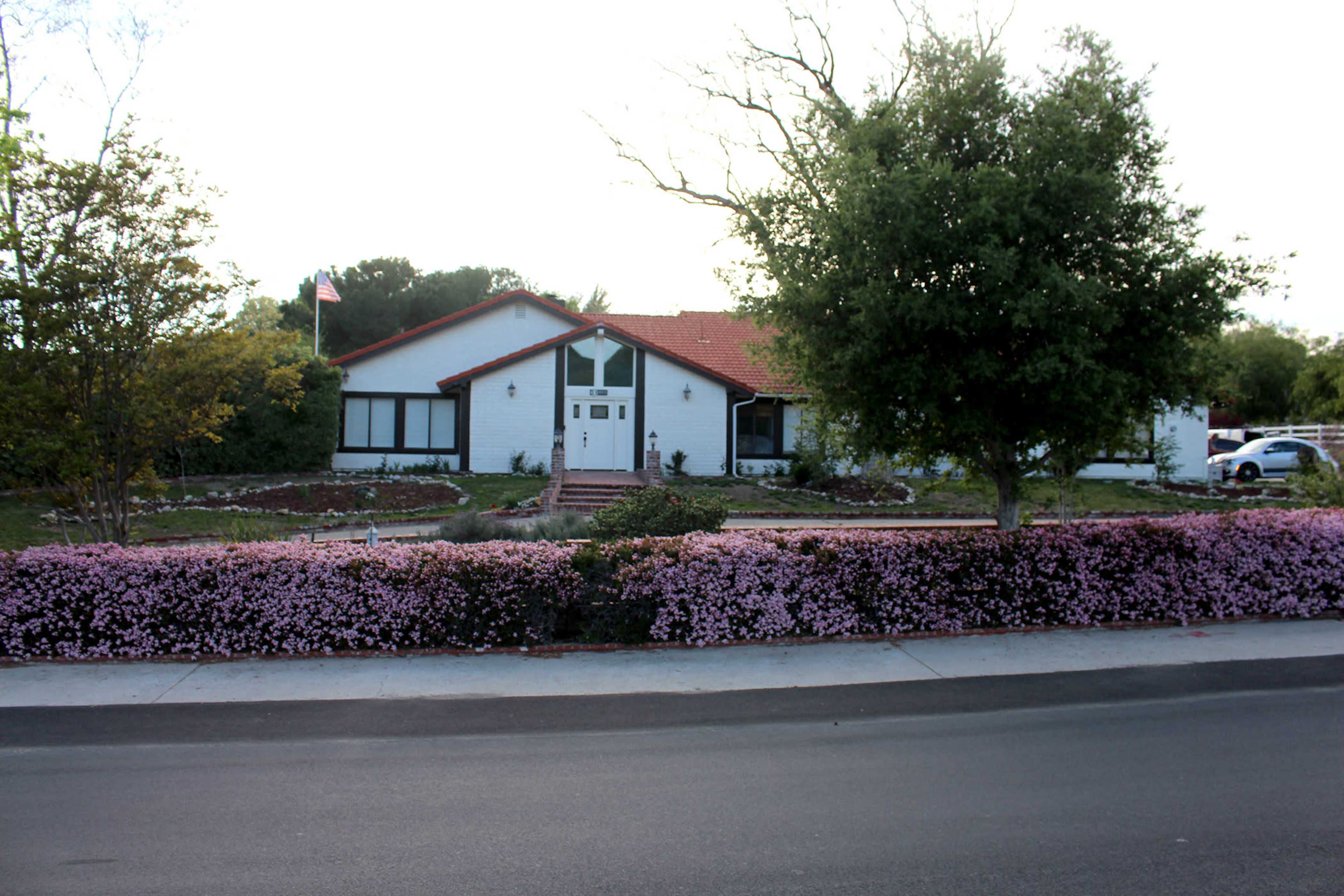 A white house with a red tiled roof is set back from the street, surrounded by a well-maintained garden featuring pink flowers along the front.