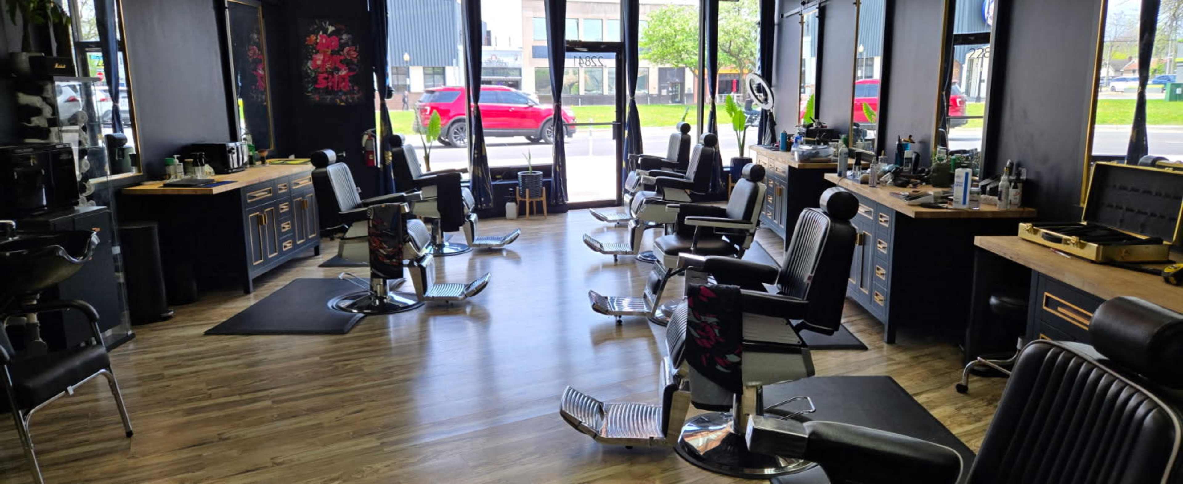 The image shows a barbershop interior with several styling chairs arranged in a row under large windows, and barber stations equipped with tools and products along the walls.