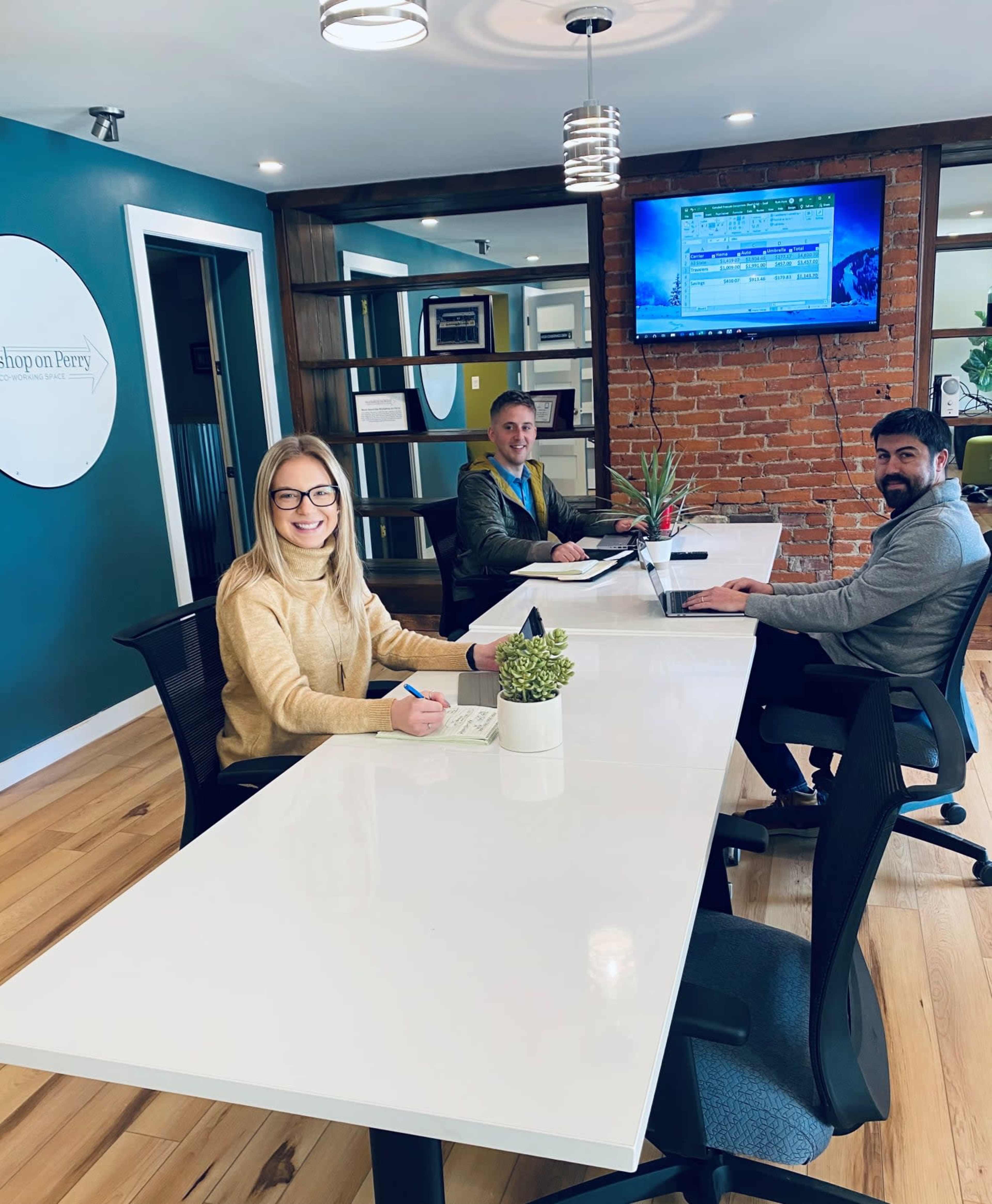 Three people are working at a modern office table with a laptop and a potted plant in a room featuring exposed brick walls and a large screen displaying information.