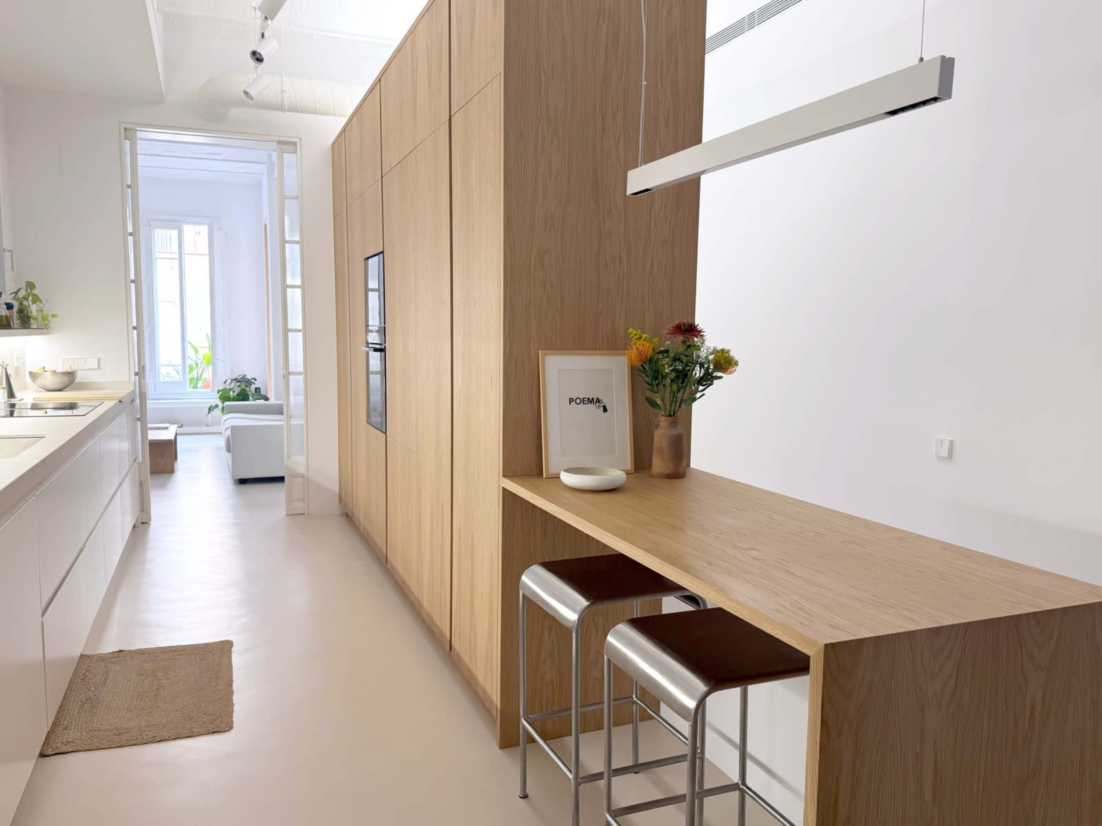 The image shows a modern kitchen with a wooden cabinetry unit, a small dining area with two stools, and a doorway leading to a well-lit living space.