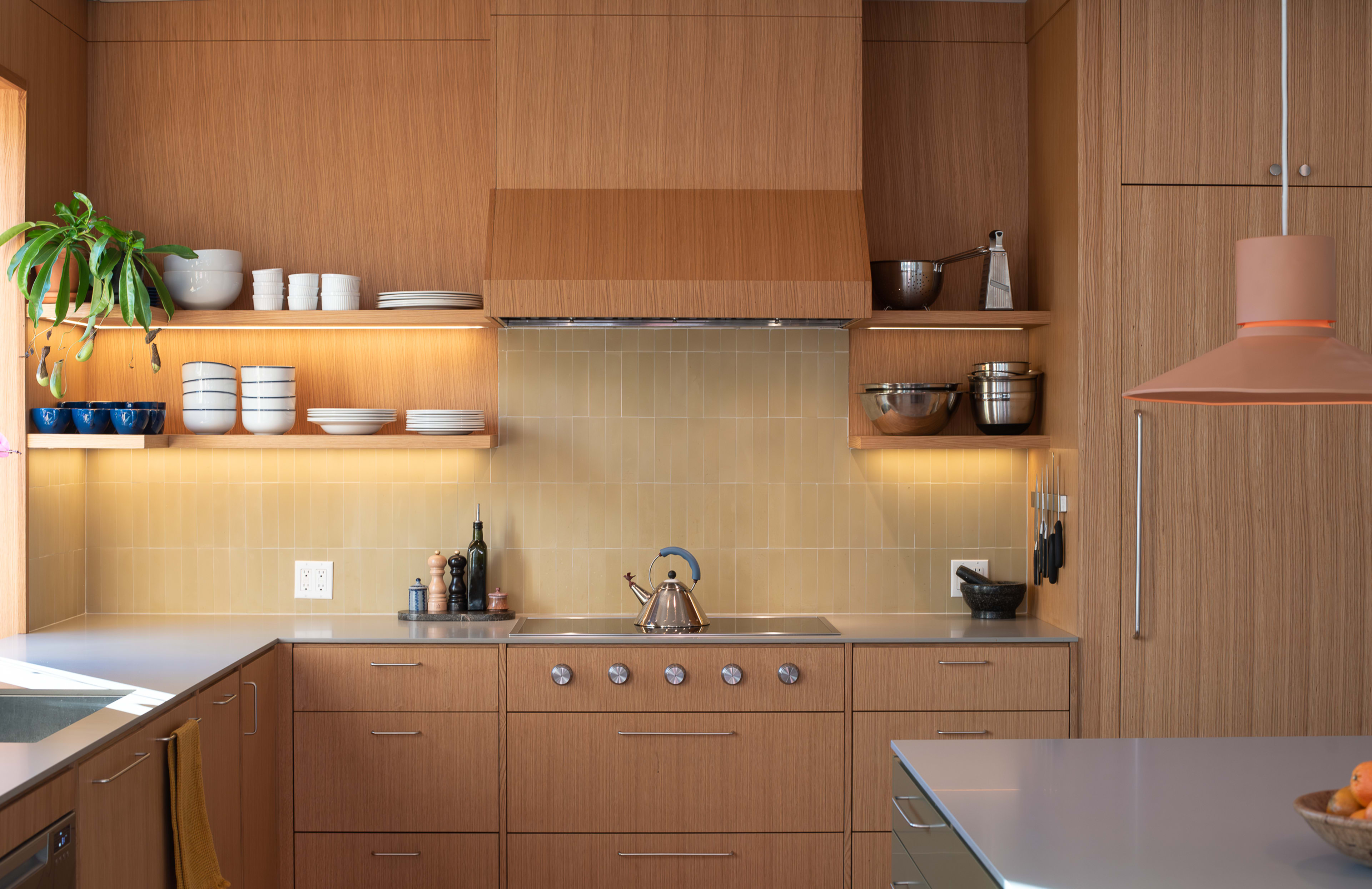 The image shows a modern kitchen with wooden cabinetry, open shelves displaying dishes, and a stove with a metallic kettle on the countertop.