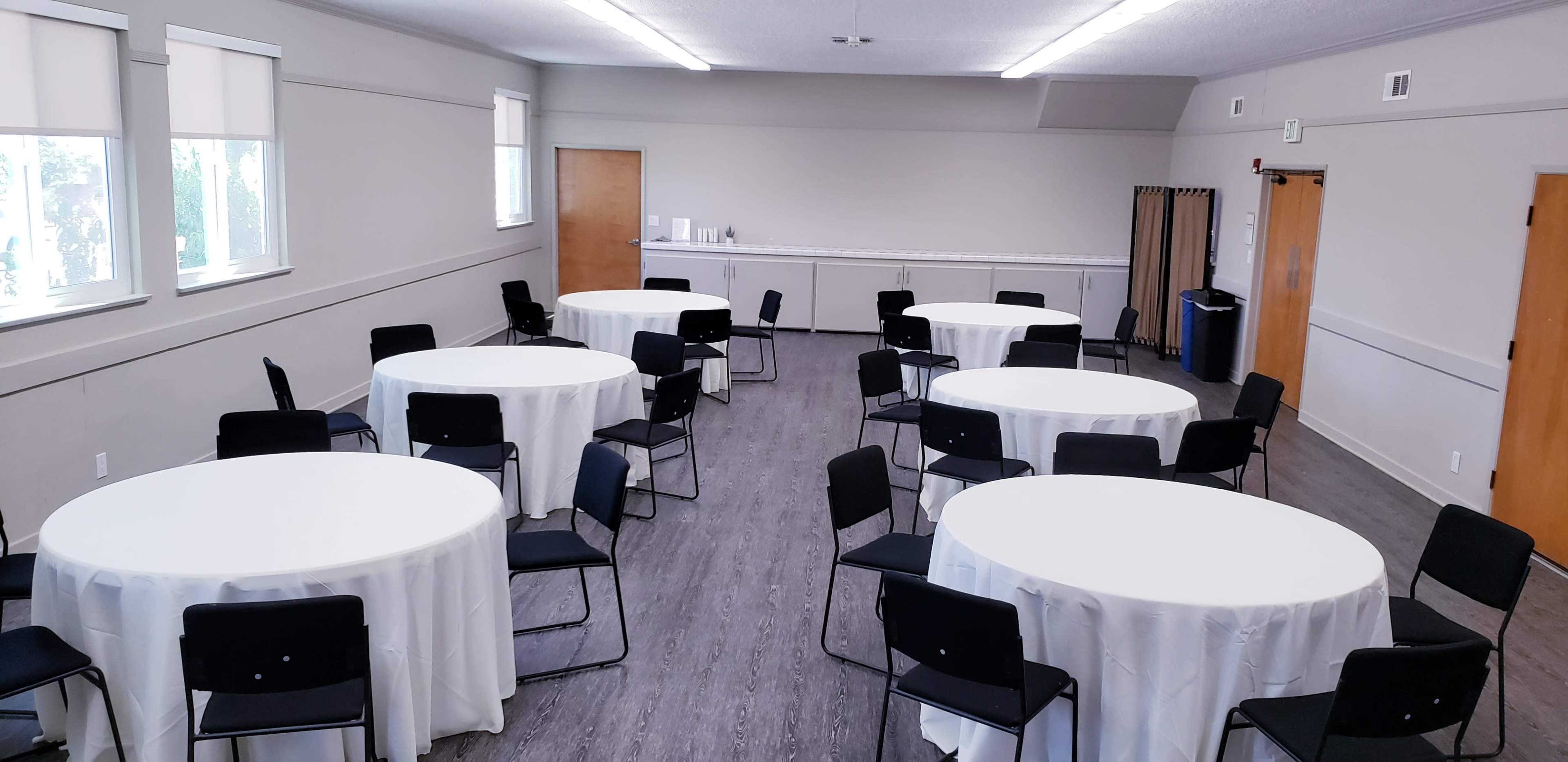 The image shows a room set up with multiple round tables covered with white tablecloths, surrounded by black chairs, and featuring plain walls and a few doors.