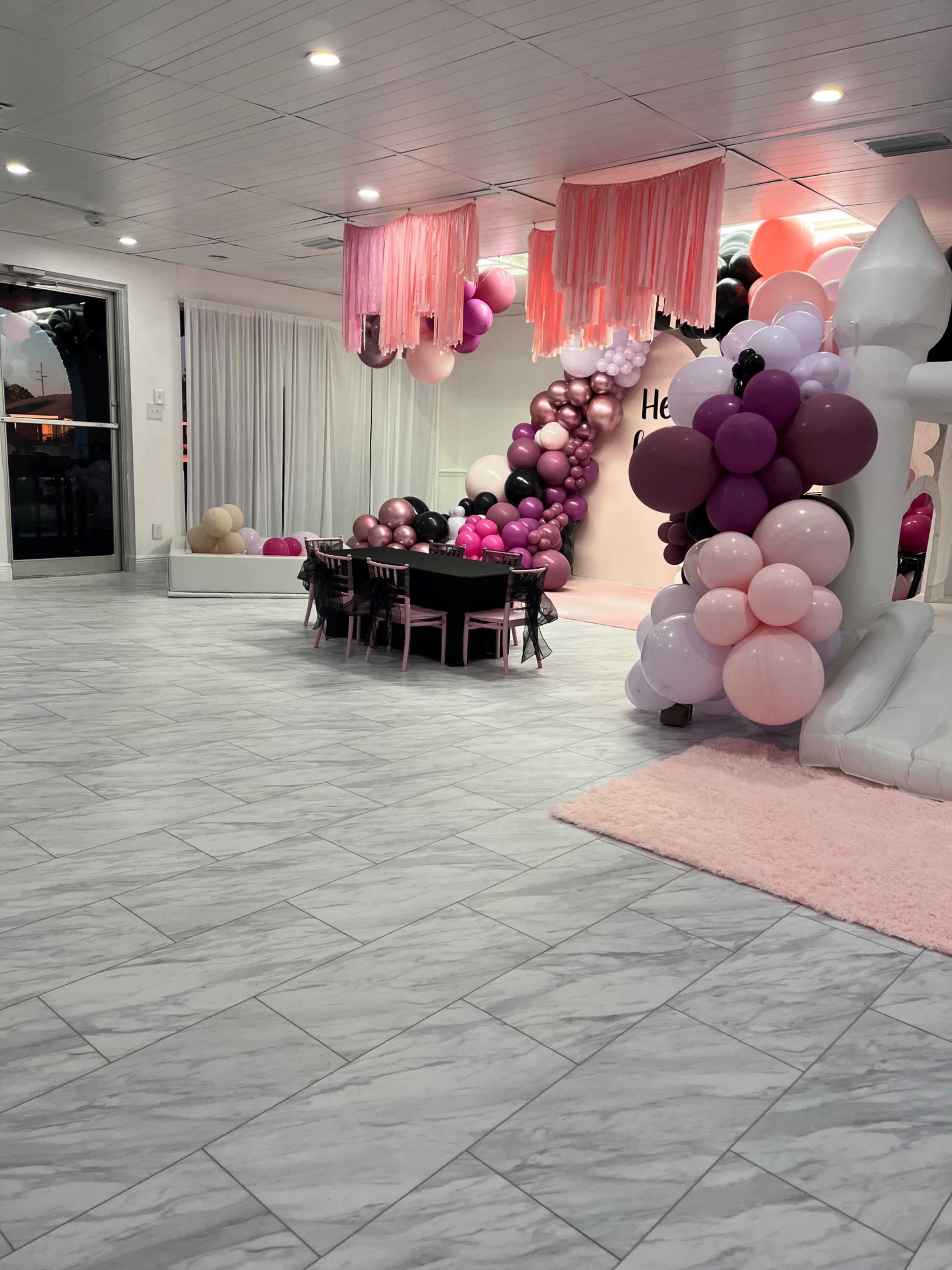 The scene features a spacious event room decorated with pink, black, and purple balloons, a table set with chairs, and a white bounce house in one corner.