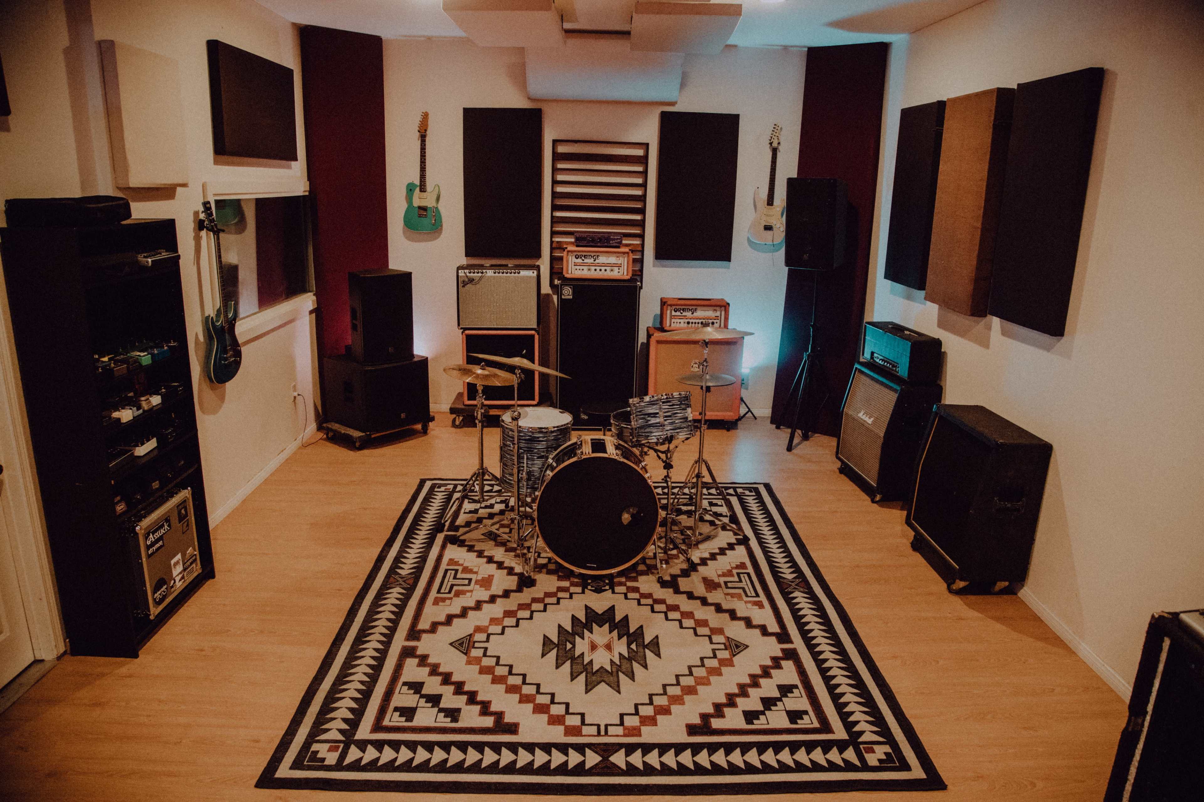 The image shows a music studio featuring a drum set in the center, surrounded by amplifiers and guitars mounted on the walls.