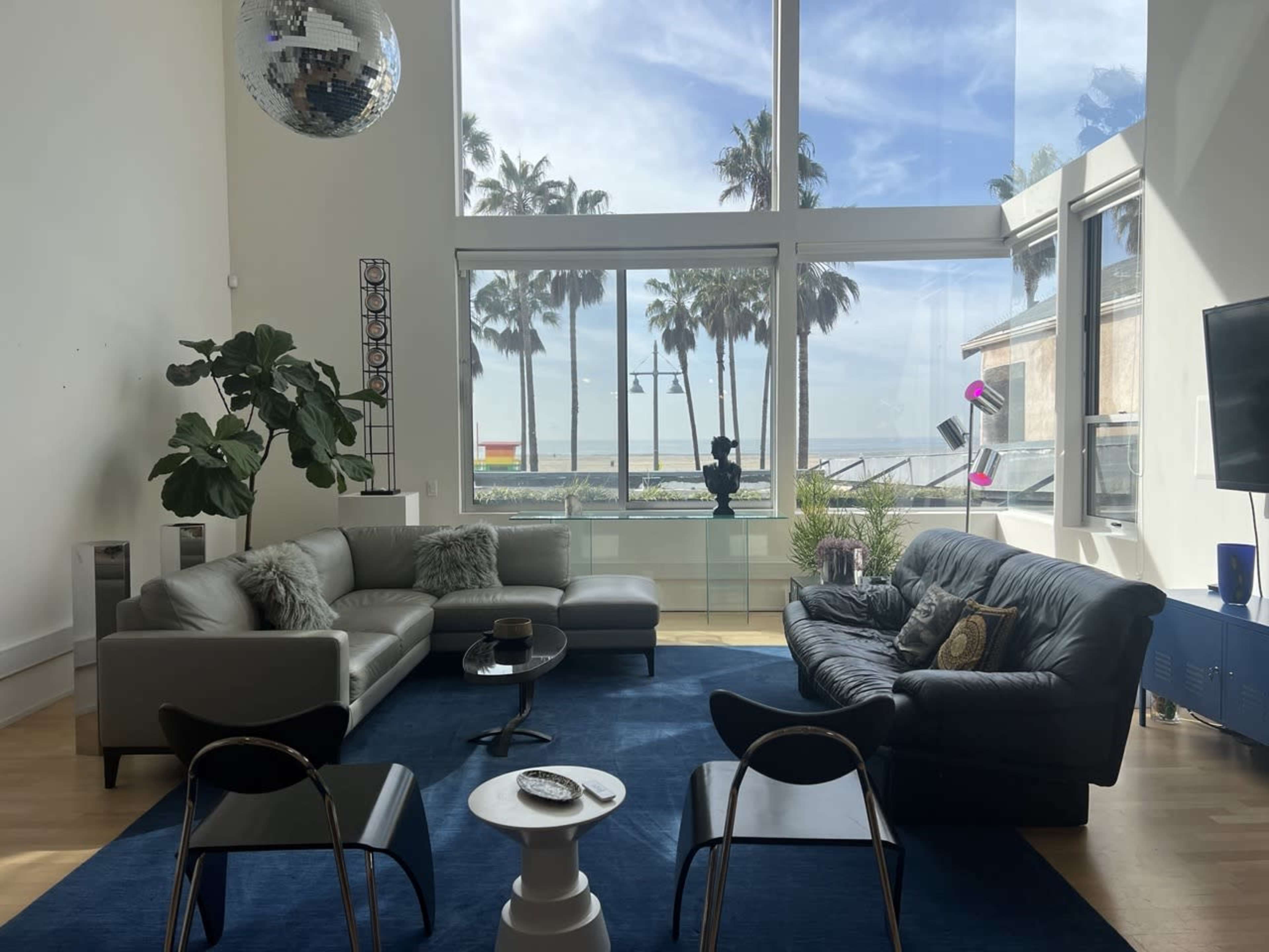 A modern living room features a large window showcasing palm trees and the ocean, with two sofas, a coffee table, and a disco ball hanging from the ceiling.