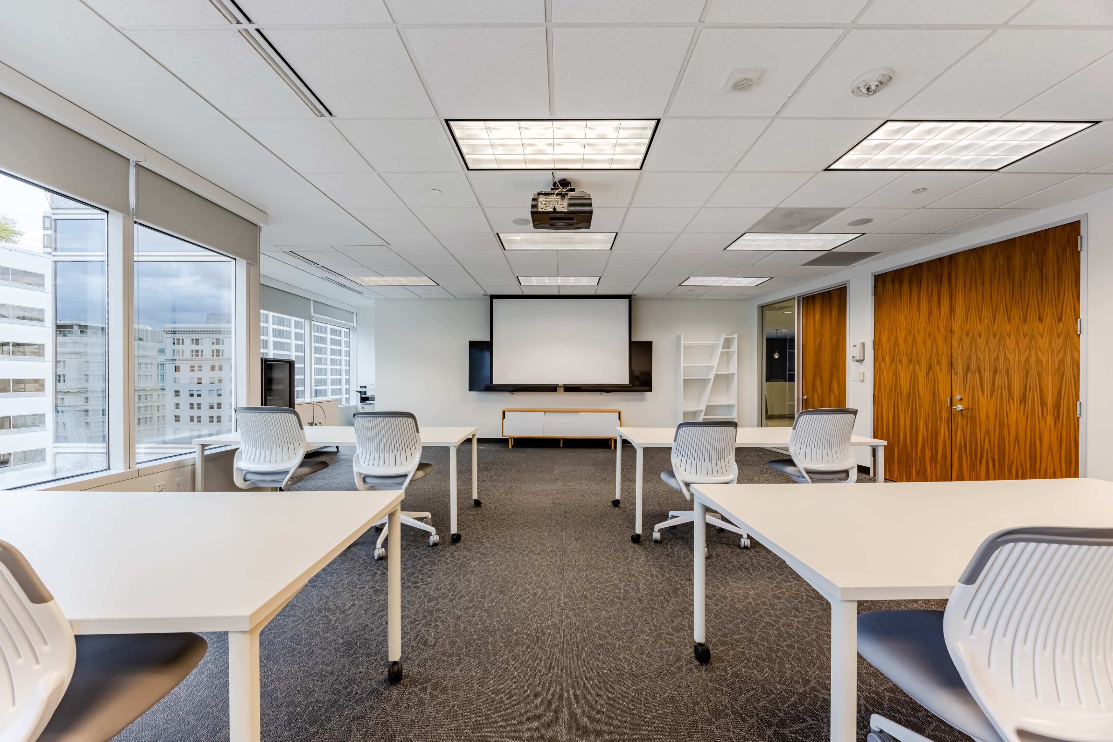 A modern conference room features several white tables arranged for meetings, a projector screen at the front, and windows providing natural light.
