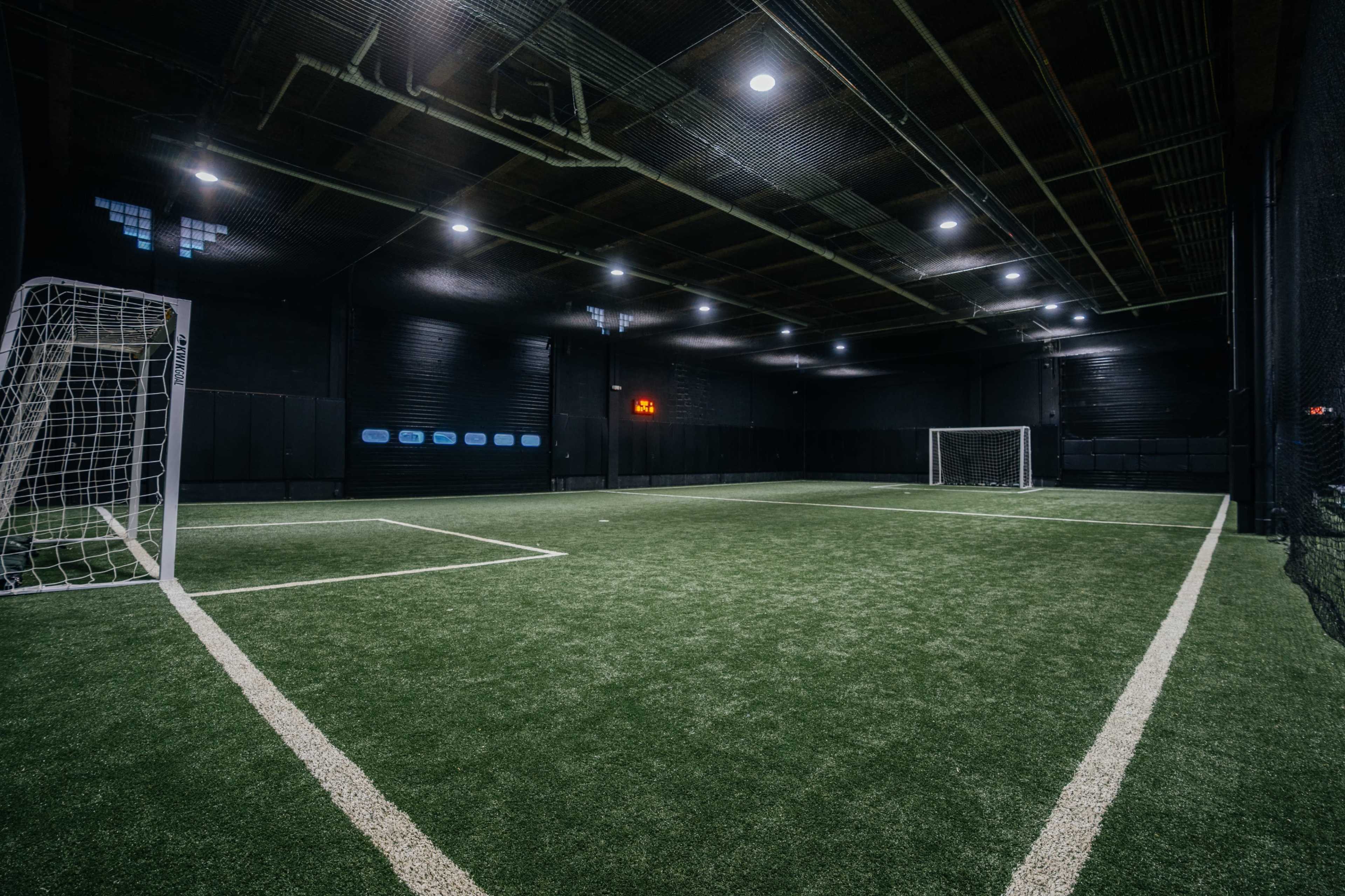 The image shows an indoor soccer field with artificial turf, featuring two goals and bright overhead lights.