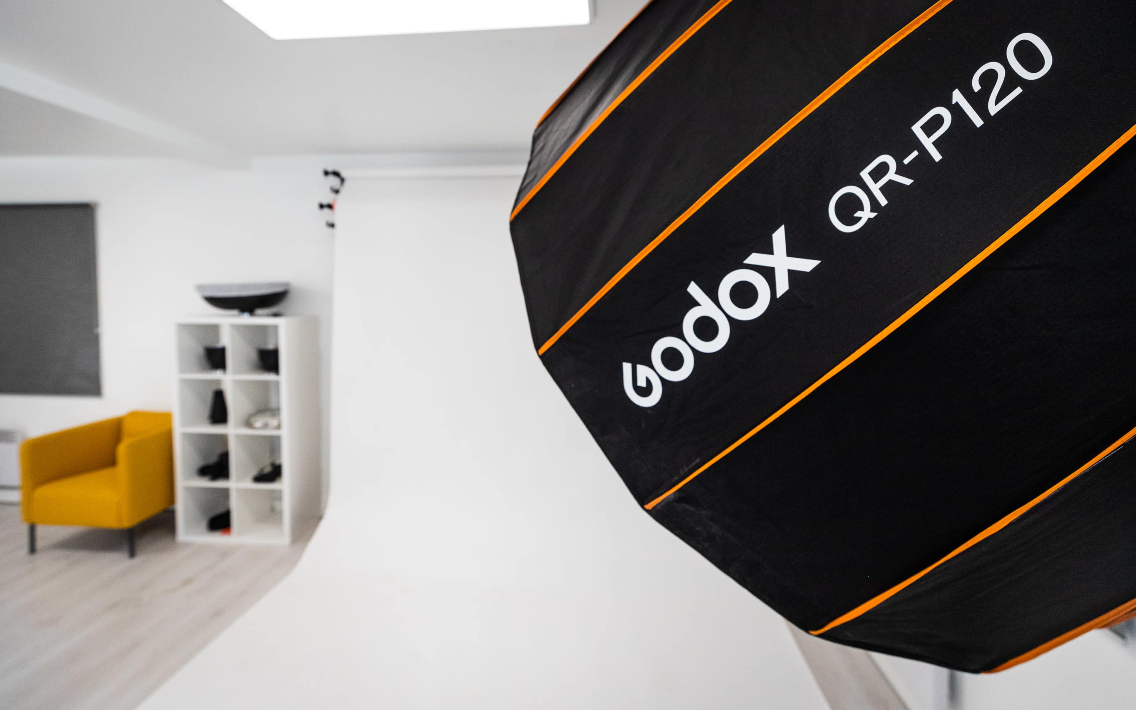 A Godox QR-P120 softbox is prominently featured in the foreground, with a minimalistic studio setup including a white backdrop and a yellow chair in the background.