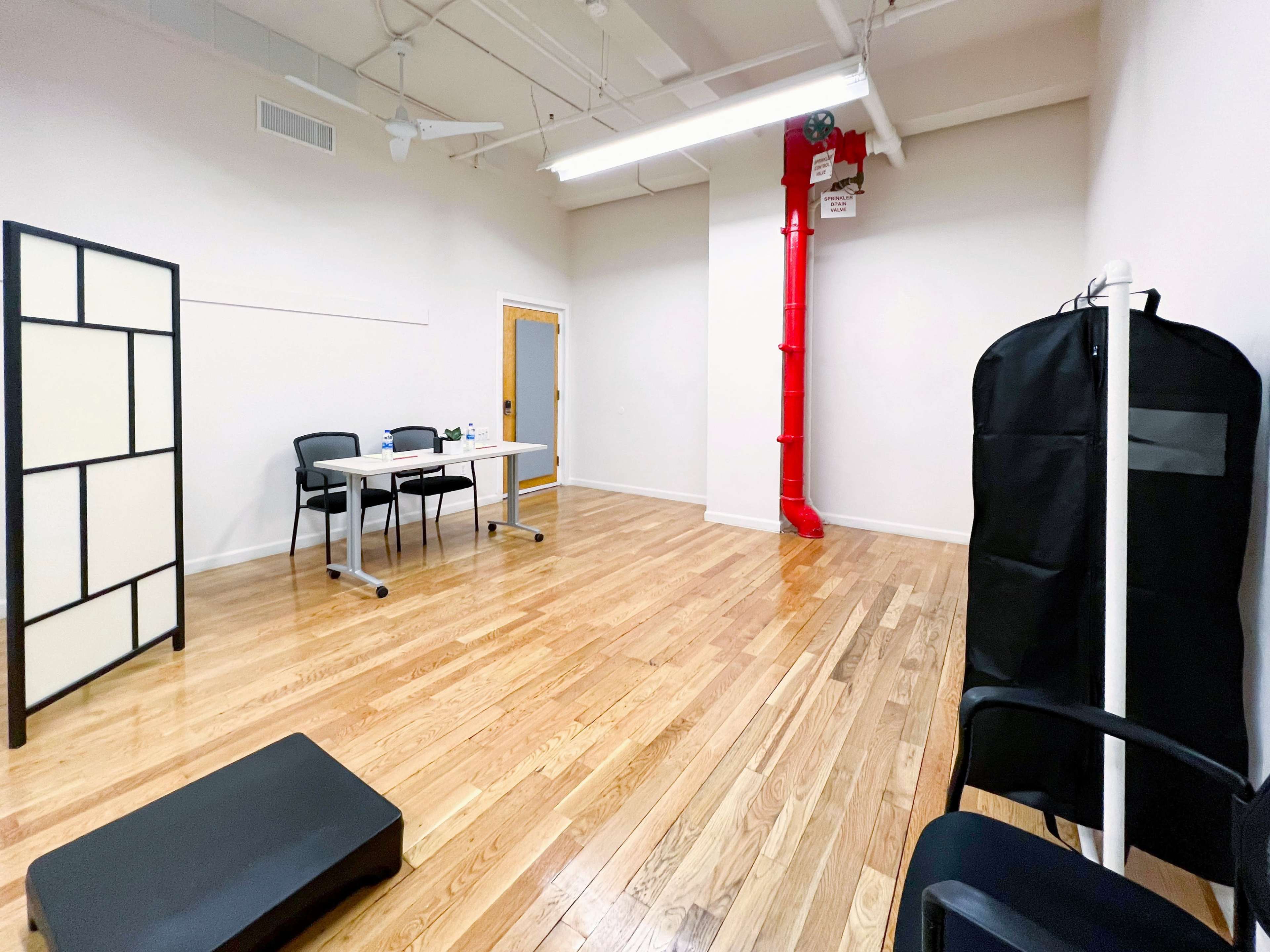 A sparse indoor room with wooden flooring, a folding screen, a table with chairs, and a red pipe on the wall.