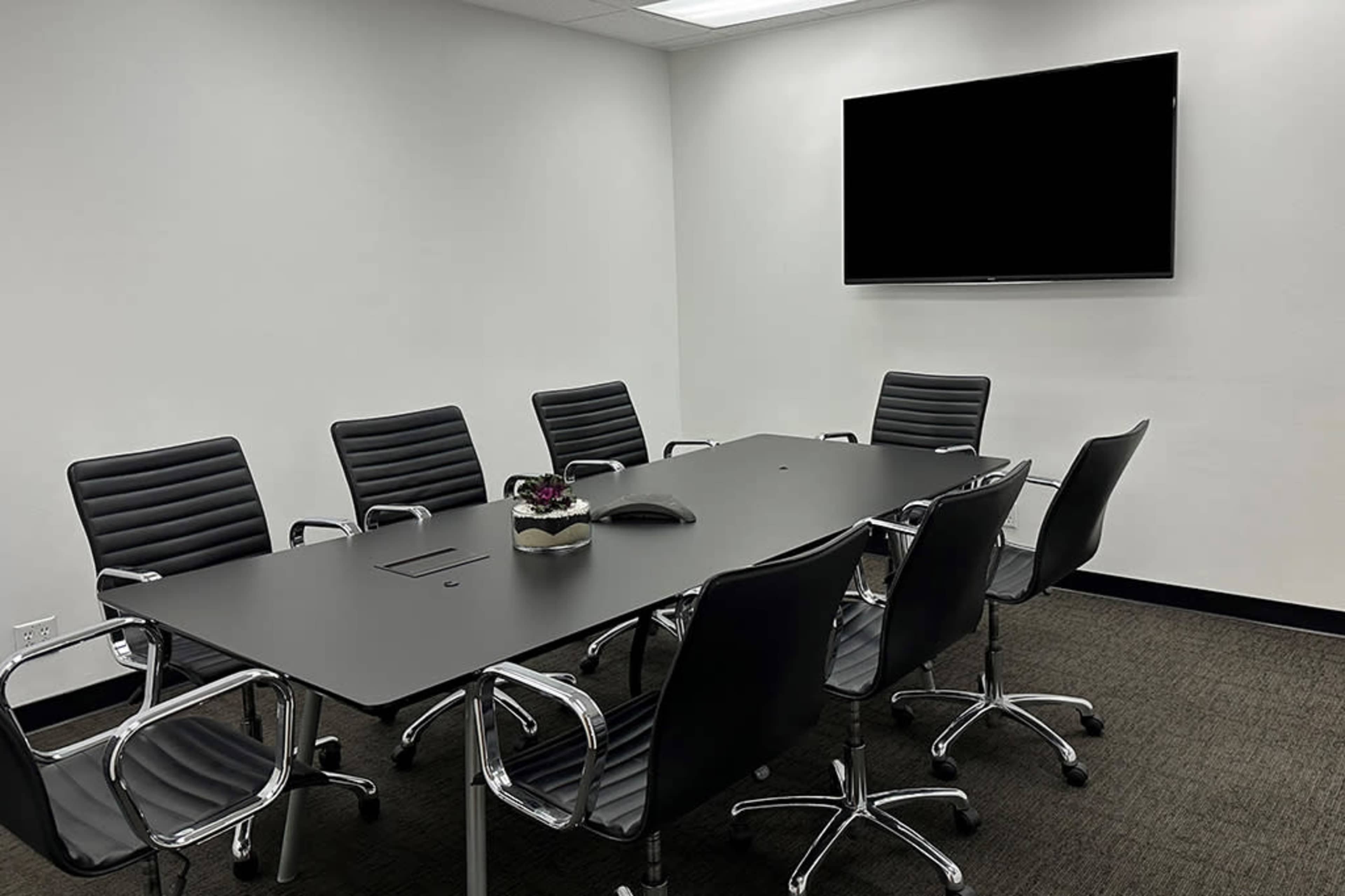 A modern conference room features a large table surrounded by sleek black chairs and a wall-mounted television.