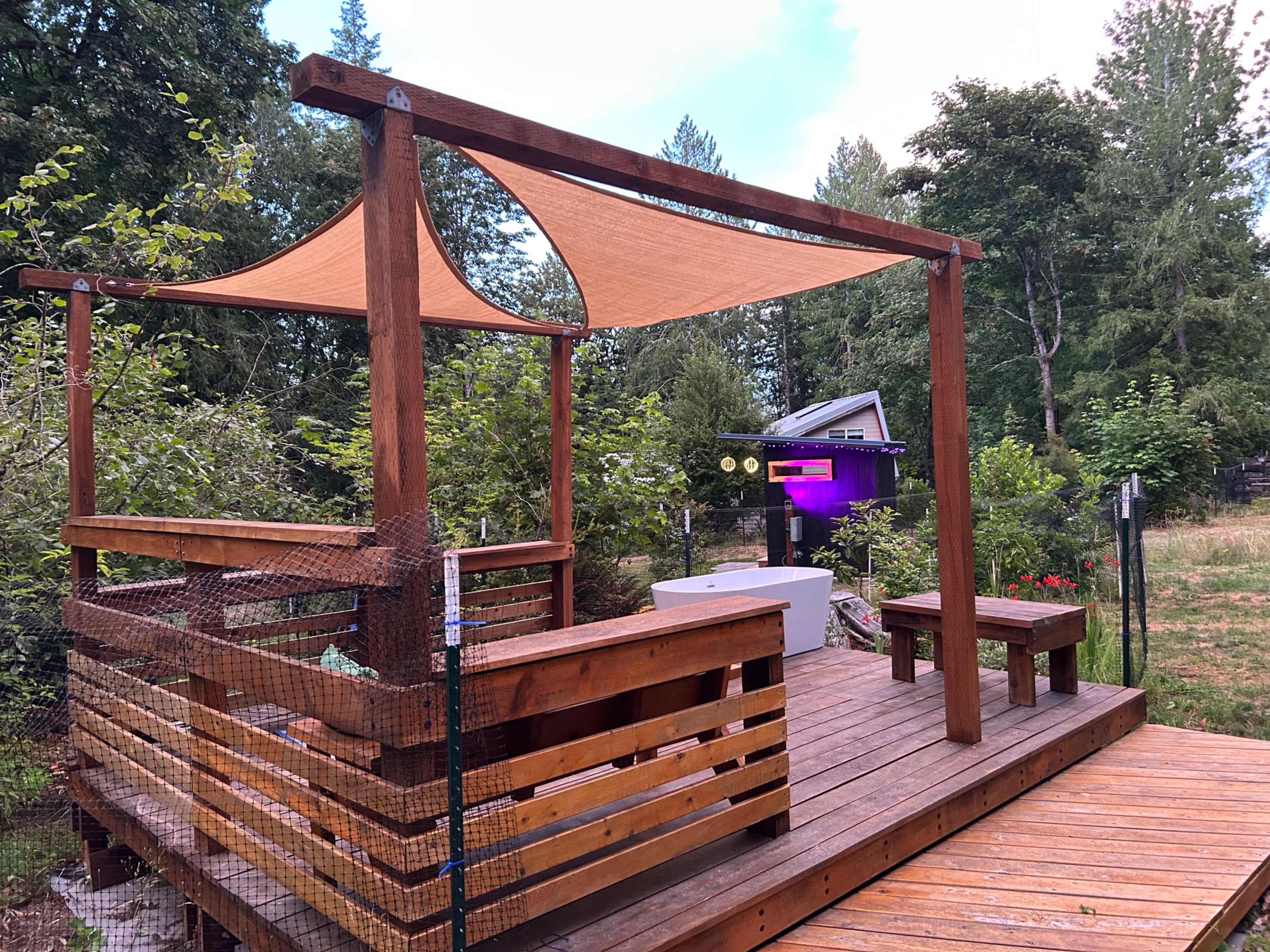 The image shows a wooden deck with a pergola featuring a fabric canopy, surrounded by greenery and a small structure in the background.