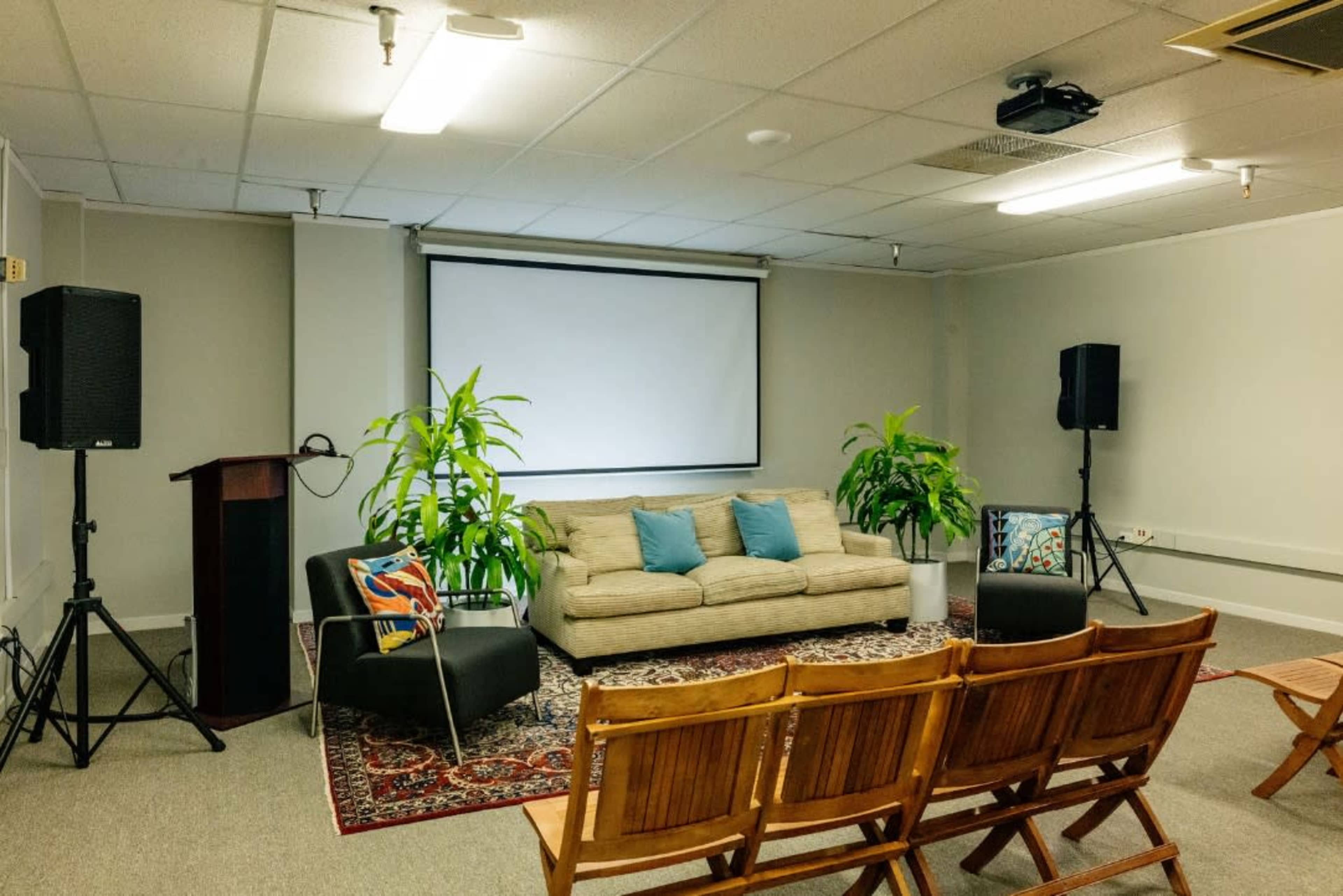 The room features a beige sofa with colorful cushions, flanked by two potted plants and a projector screen, surrounded by wooden chairs arranged in a semi-circle.