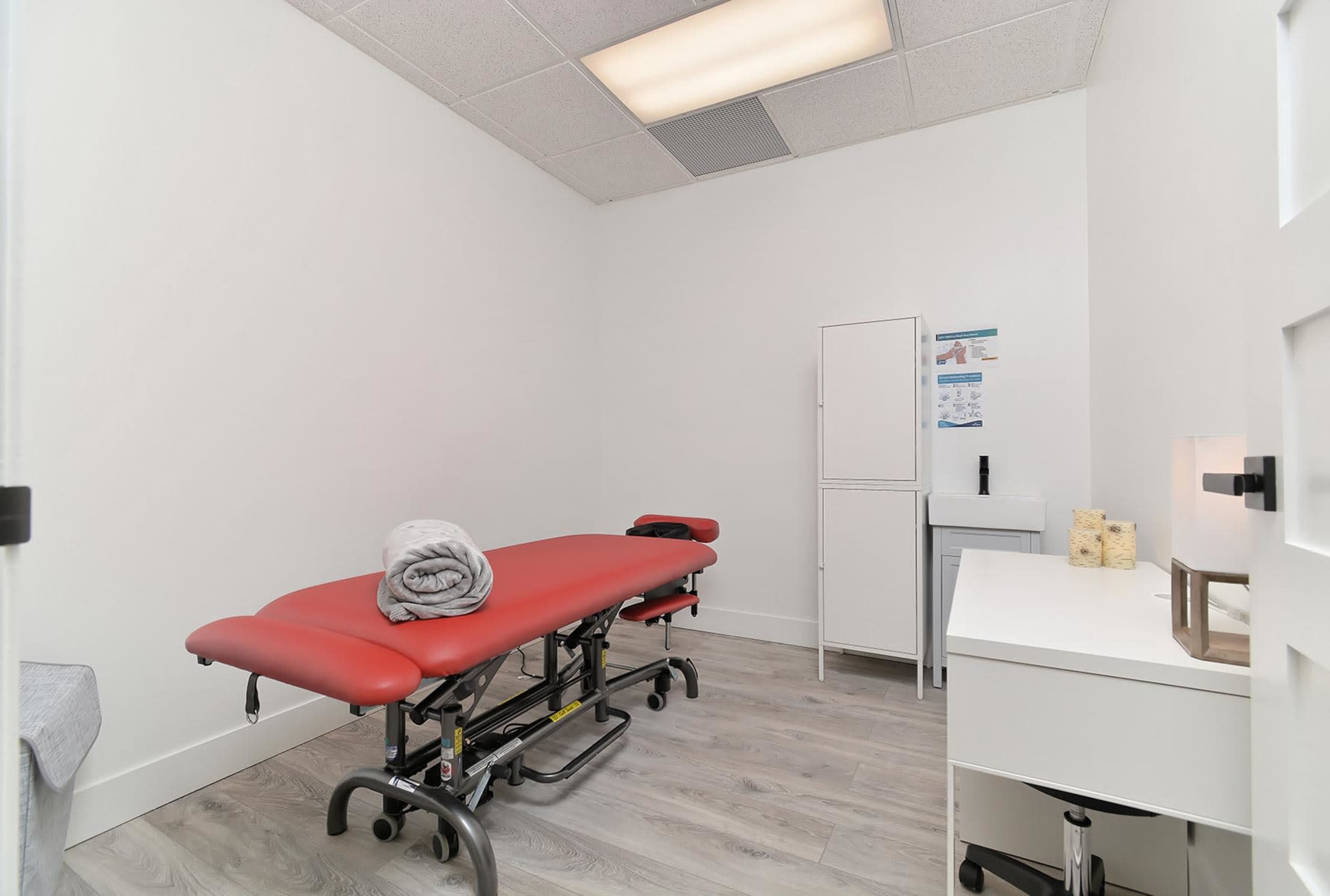 A treatment room with a red massage table, a small white cabinet, and a refrigerator against the wall.