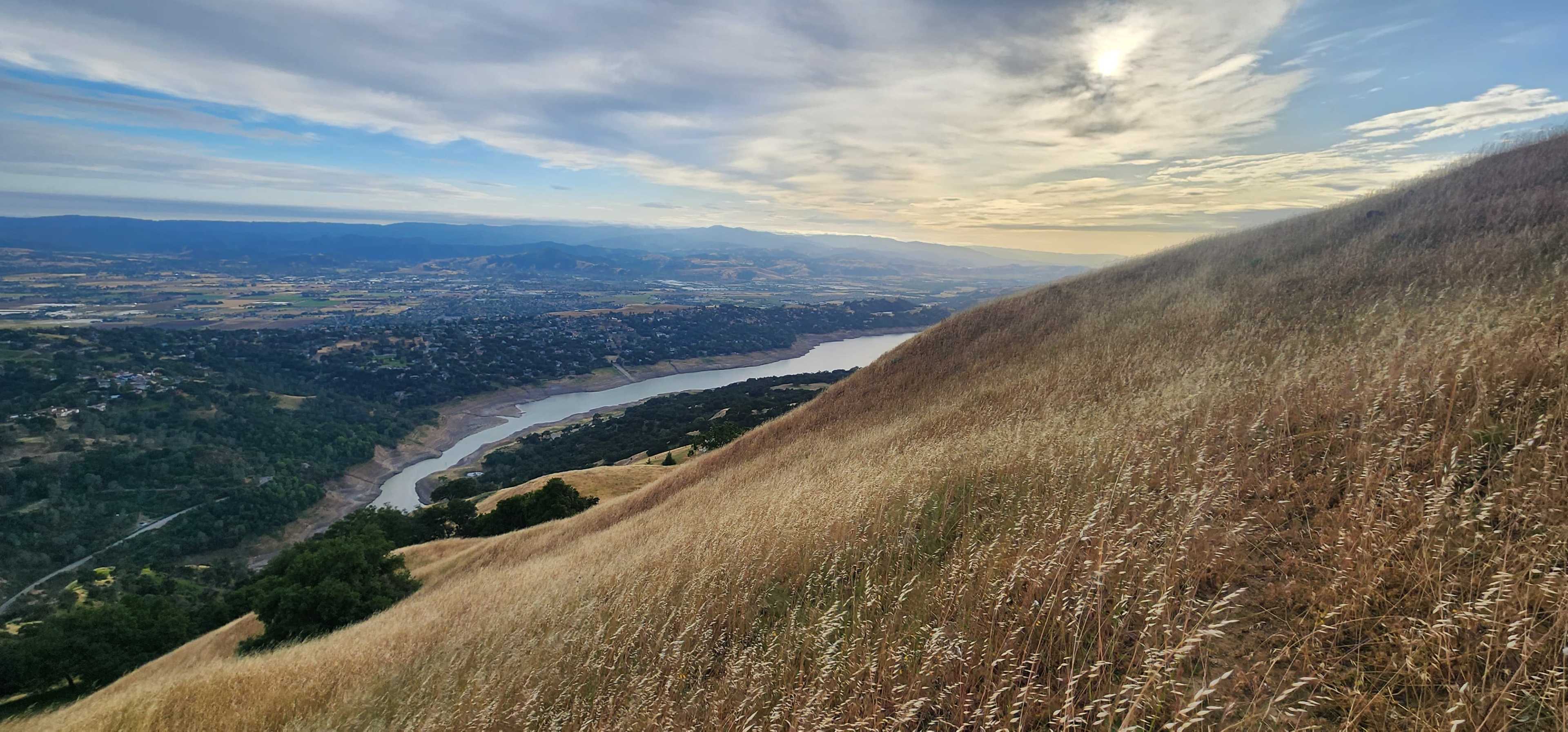 The image shows a sweeping view of a river winding through a valley, bordered by rolling hills covered in tall grass under a cloudy sky.