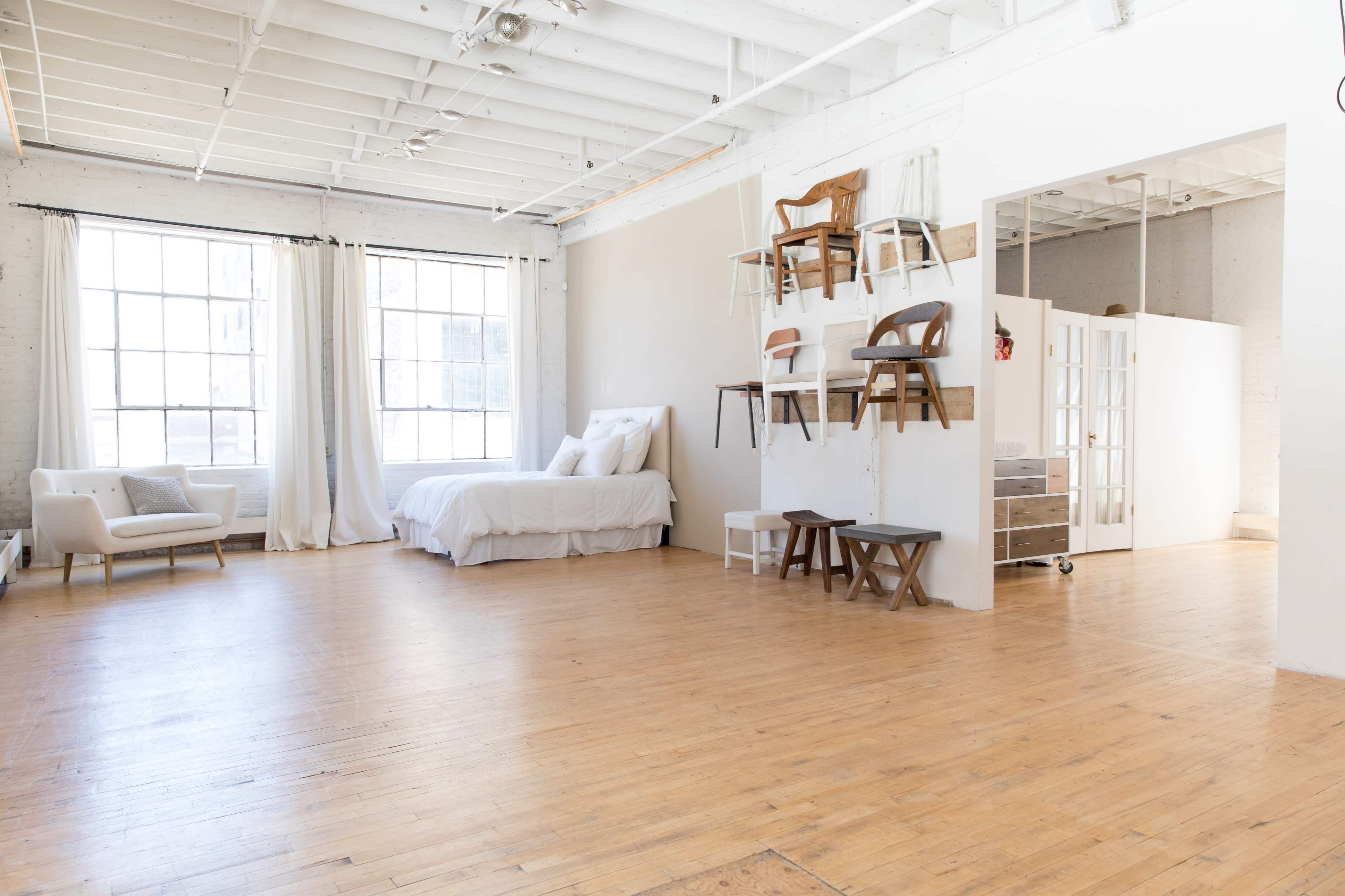 The image shows a bright, open living space featuring a bed, a seating area, and shelves with various chairs against a white wall.