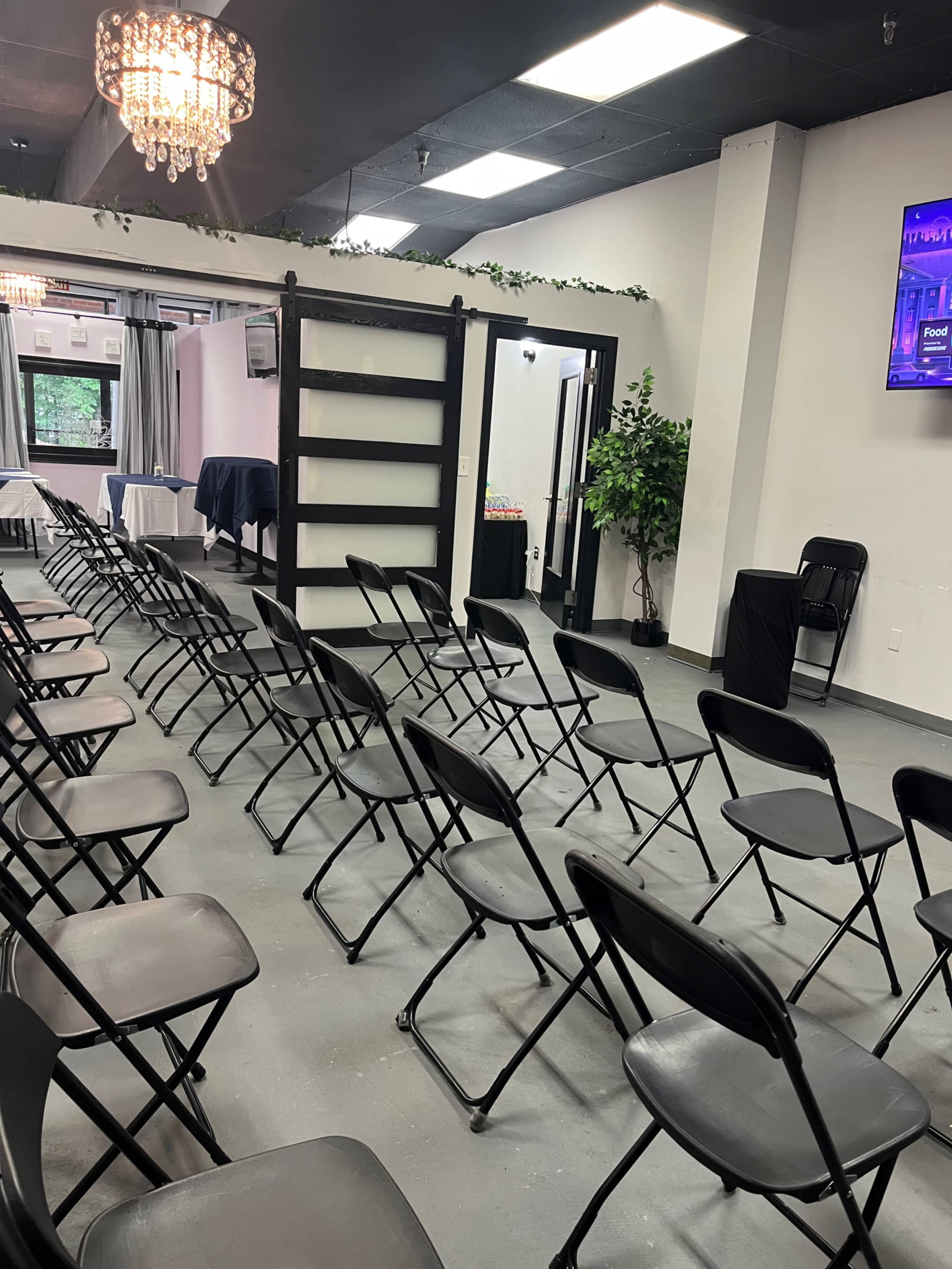 The image shows a room set up for an event, featuring rows of black folding chairs facing a wall with a television screen and decorative elements.