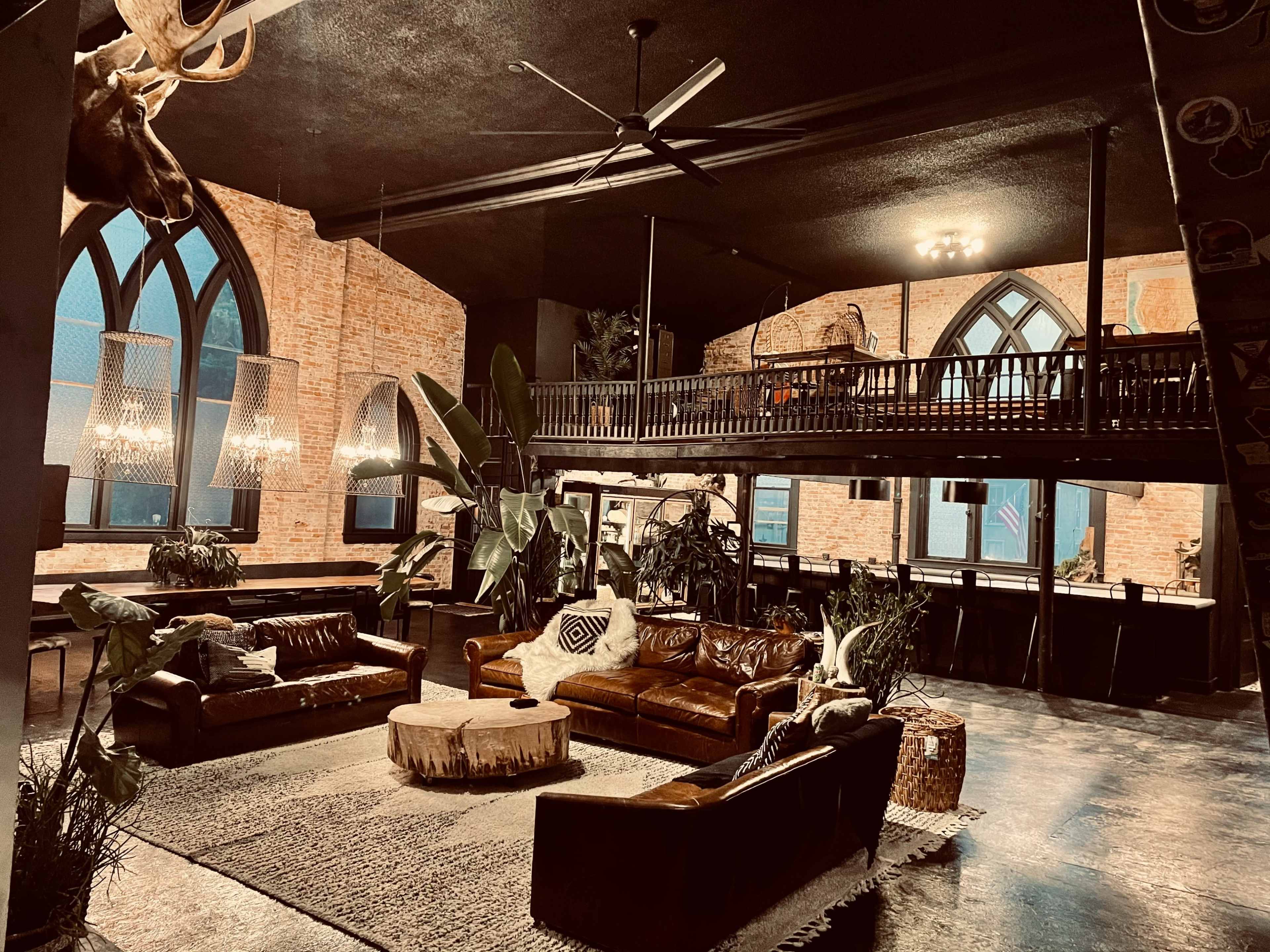 A spacious, industrial-style interior with two leather sofas, a round coffee table, large plants, and an upper loft area featuring gothic-style windows.