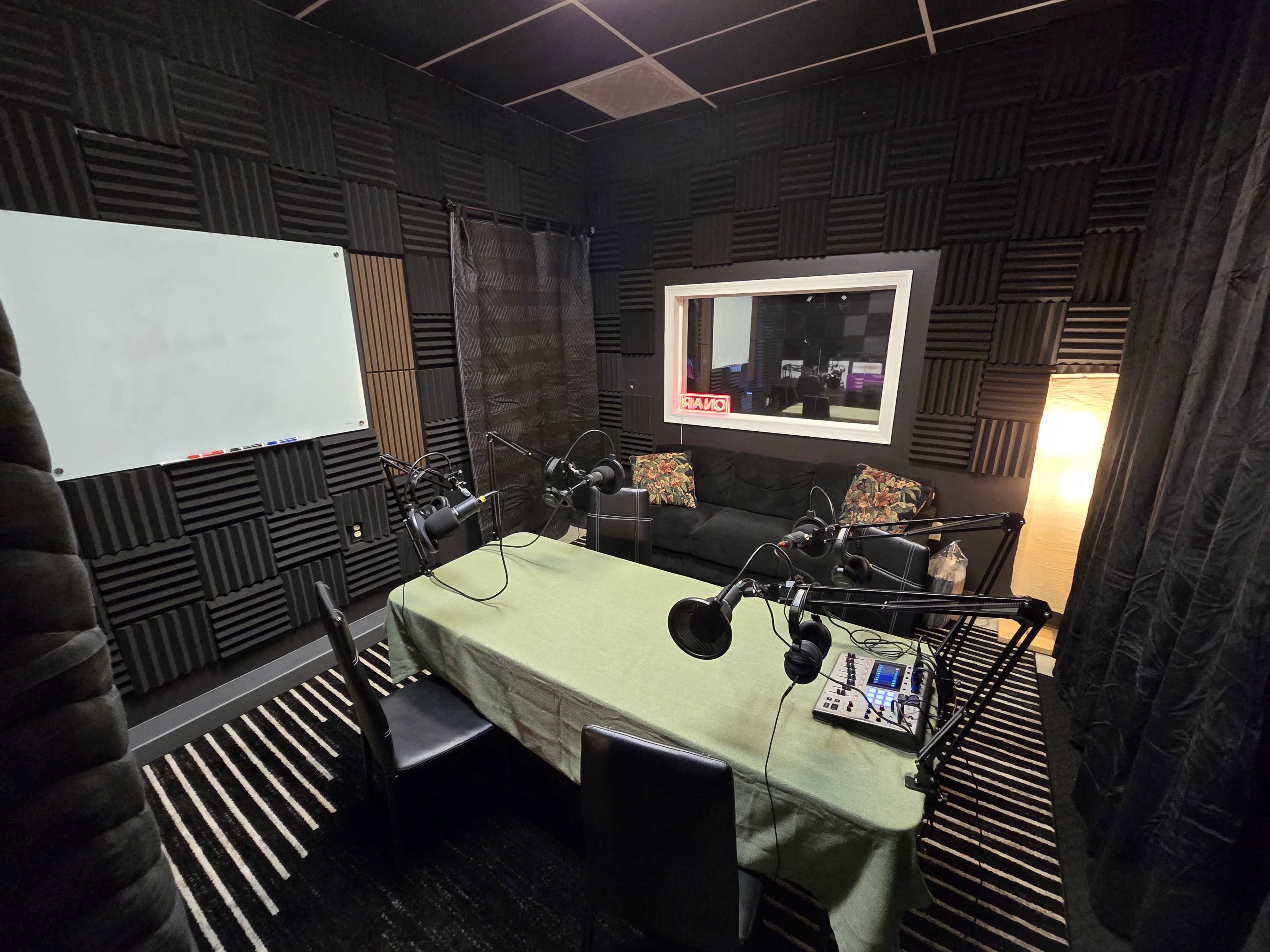 The image shows a sound studio with acoustic foam panels, a table covered with a green cloth, and multiple microphones set up on stands.