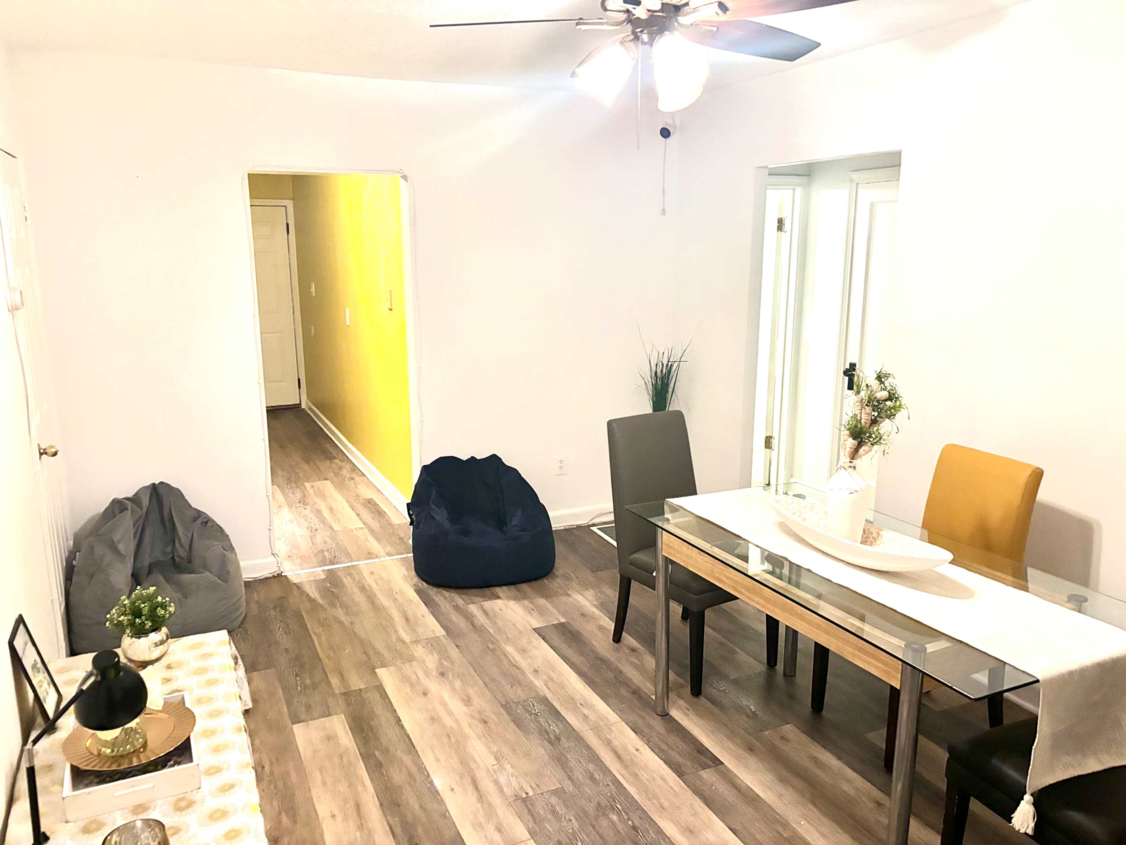 The image shows a simple, well-lit dining area featuring a glass table with four chairs, a couple of bean bags in the corner, and white walls accented by a yellow wall in an adjacent room.