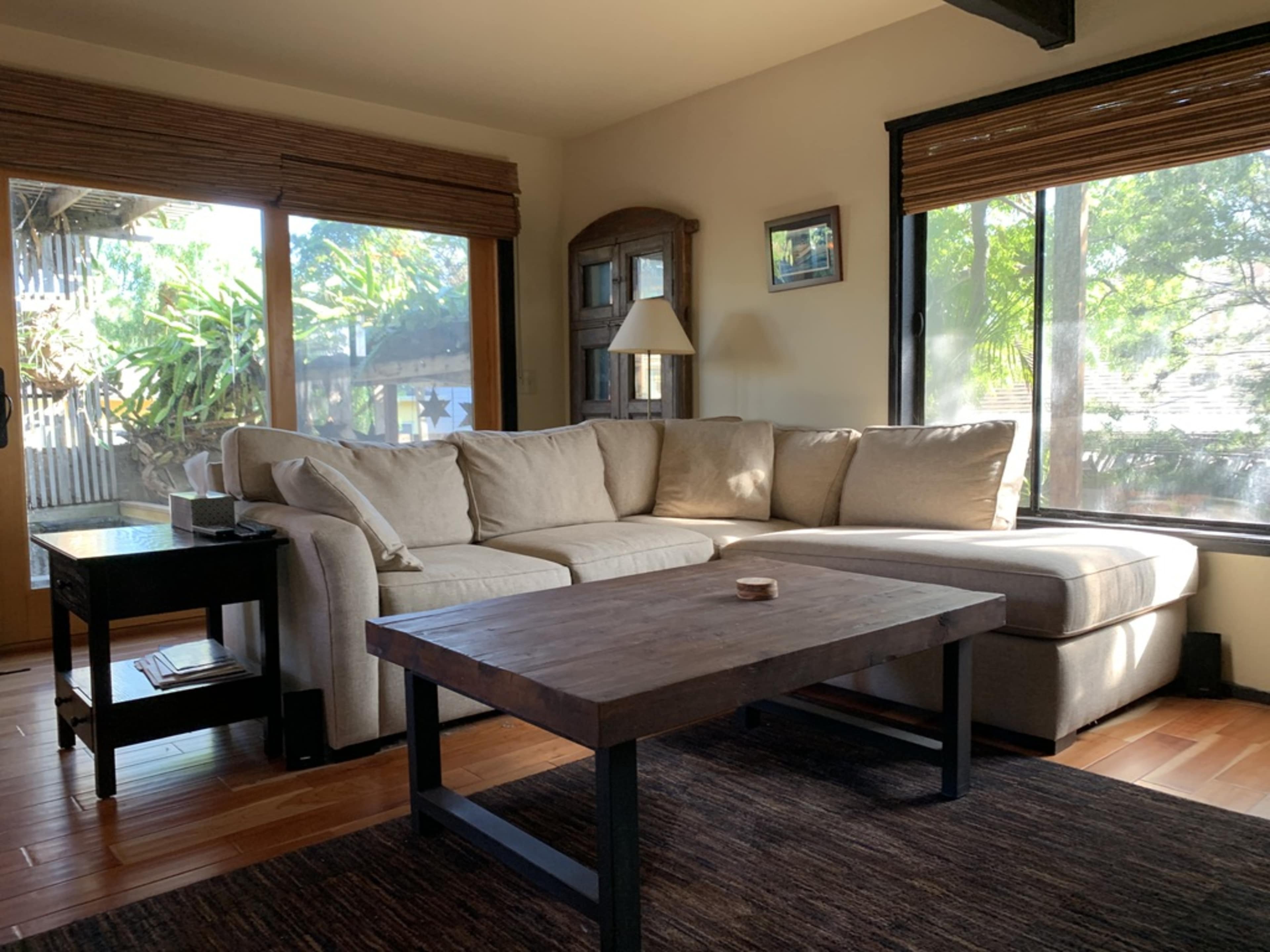 A well-lit living room features a large sectional sofa, a wooden coffee table, and large windows that showcase greenery outside.