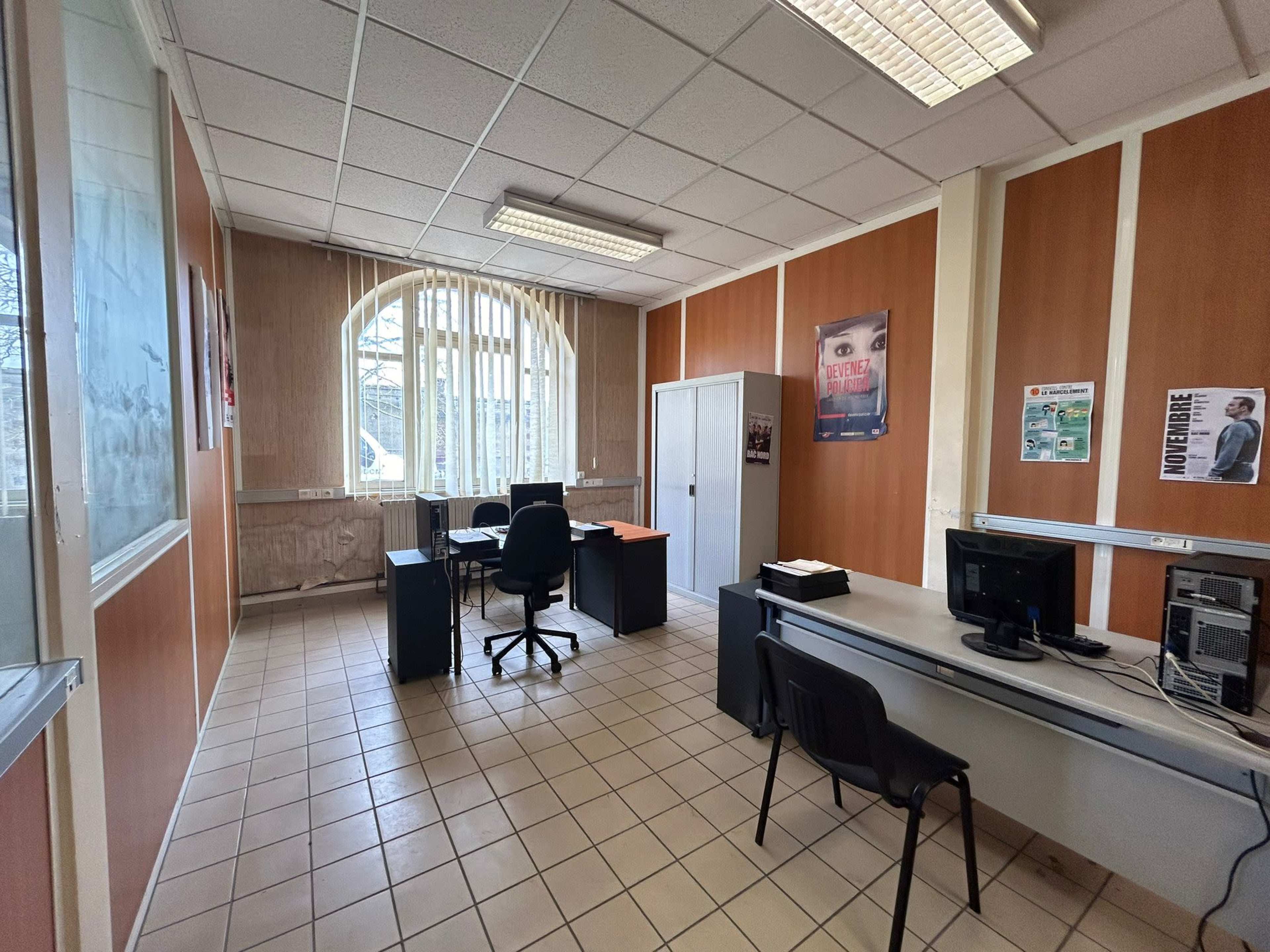 A vacant office space featuring desks, black chairs, and a computer setup against walls of wood paneling and windows.