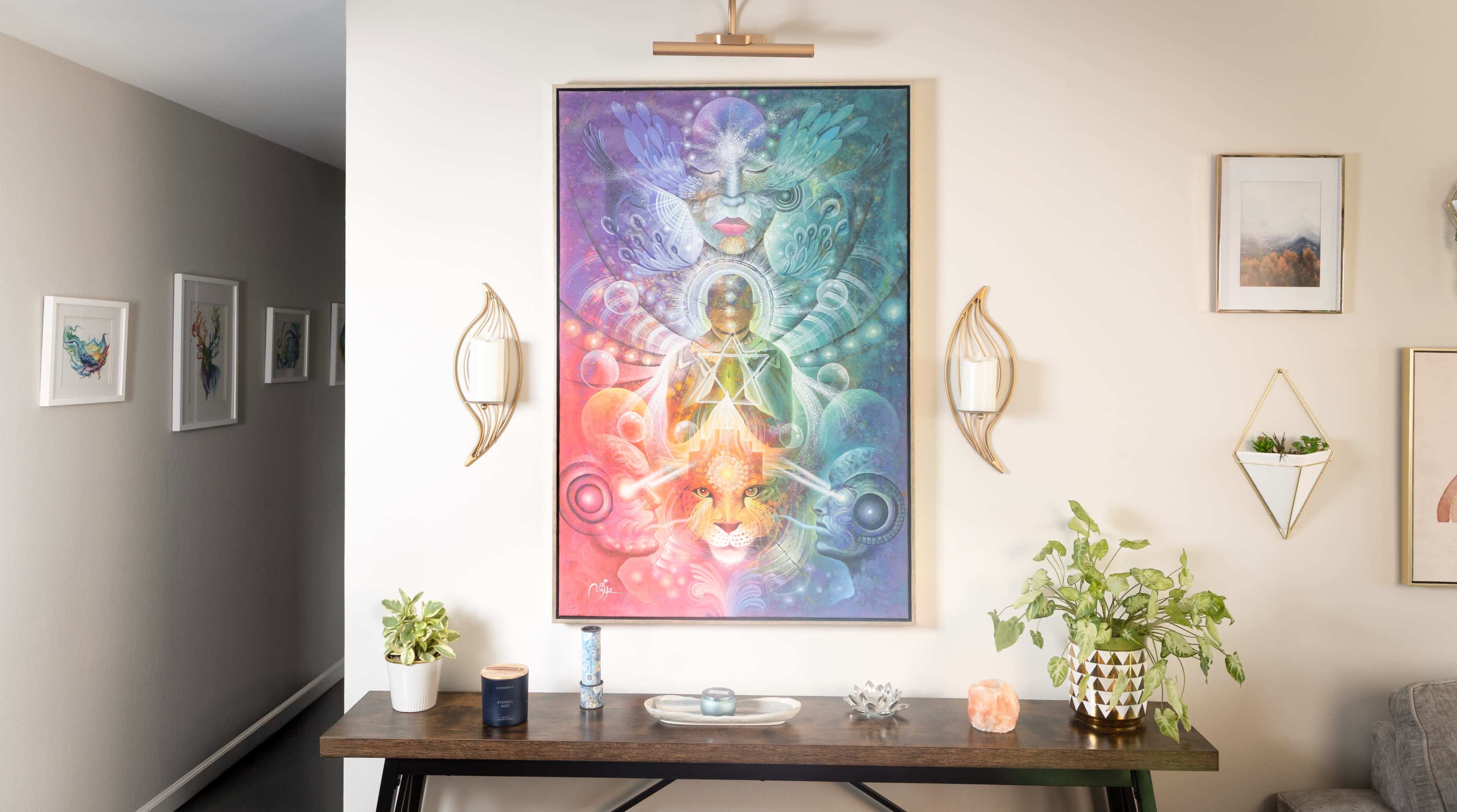 The image shows a colorful artwork featuring a figure surrounded by geometric patterns and animals, displayed on a wall above a wooden table decorated with plants and candles.