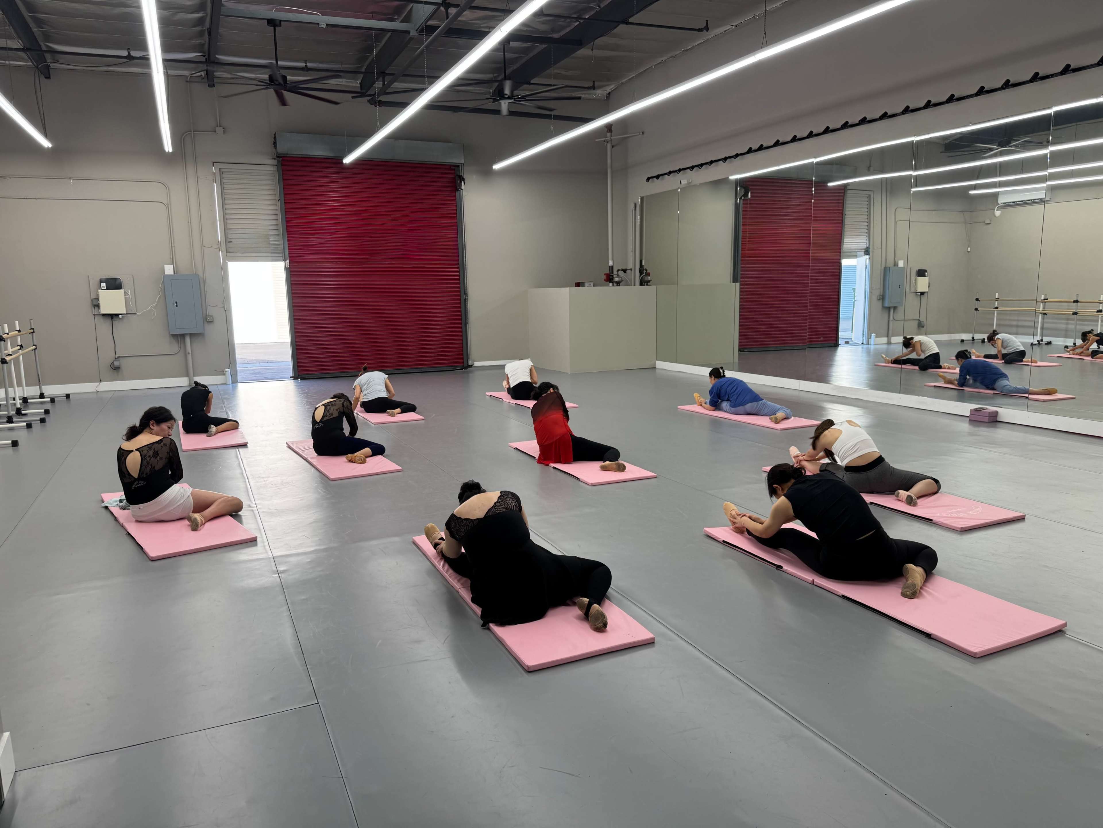 A group of people are practicing yoga on pink mats in a spacious studio with mirrors and red walls.