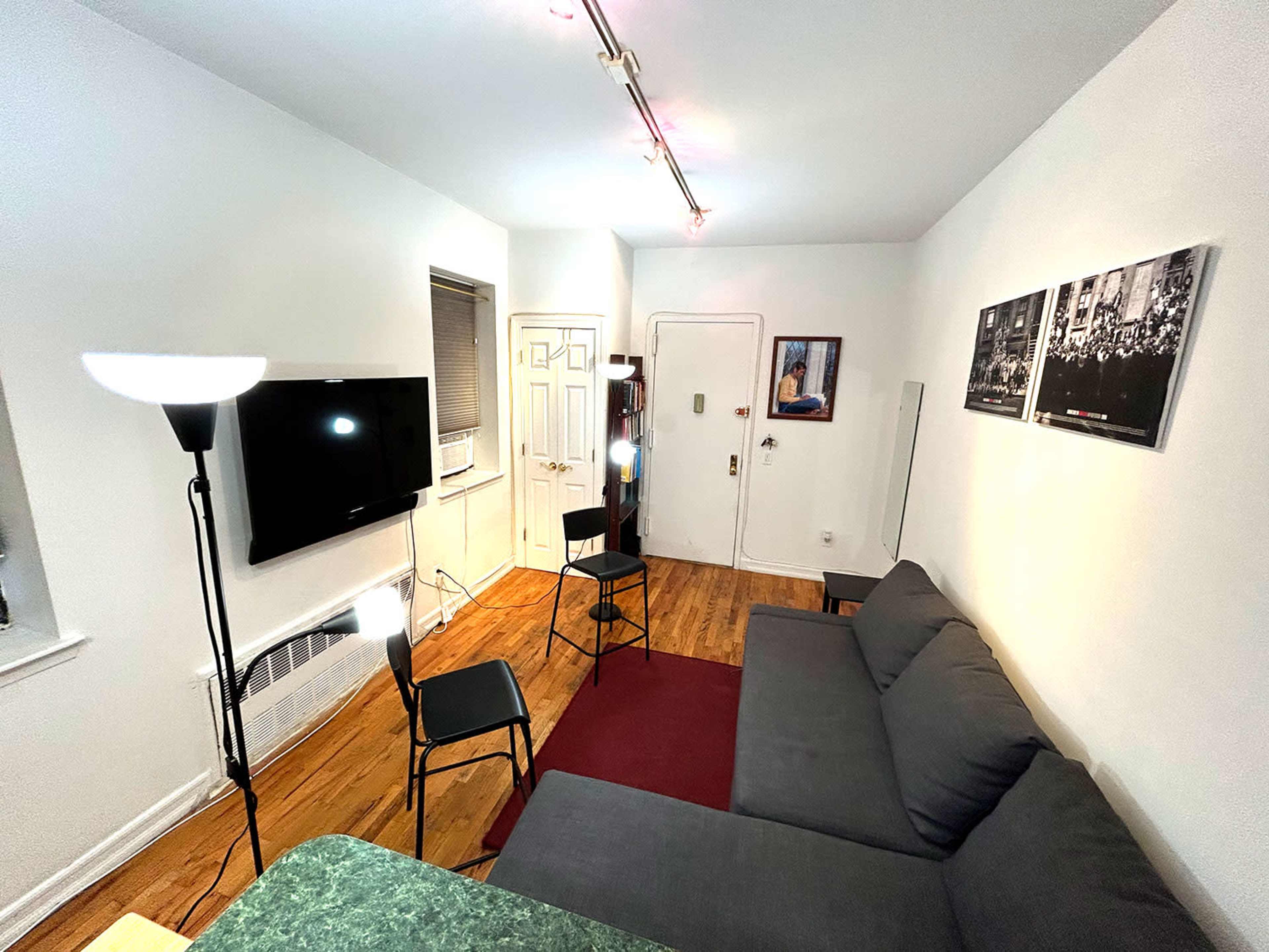 A small living room features a gray sofa, a television mounted on the wall, two chairs positioned around a green table, and bright lighting fixtures.