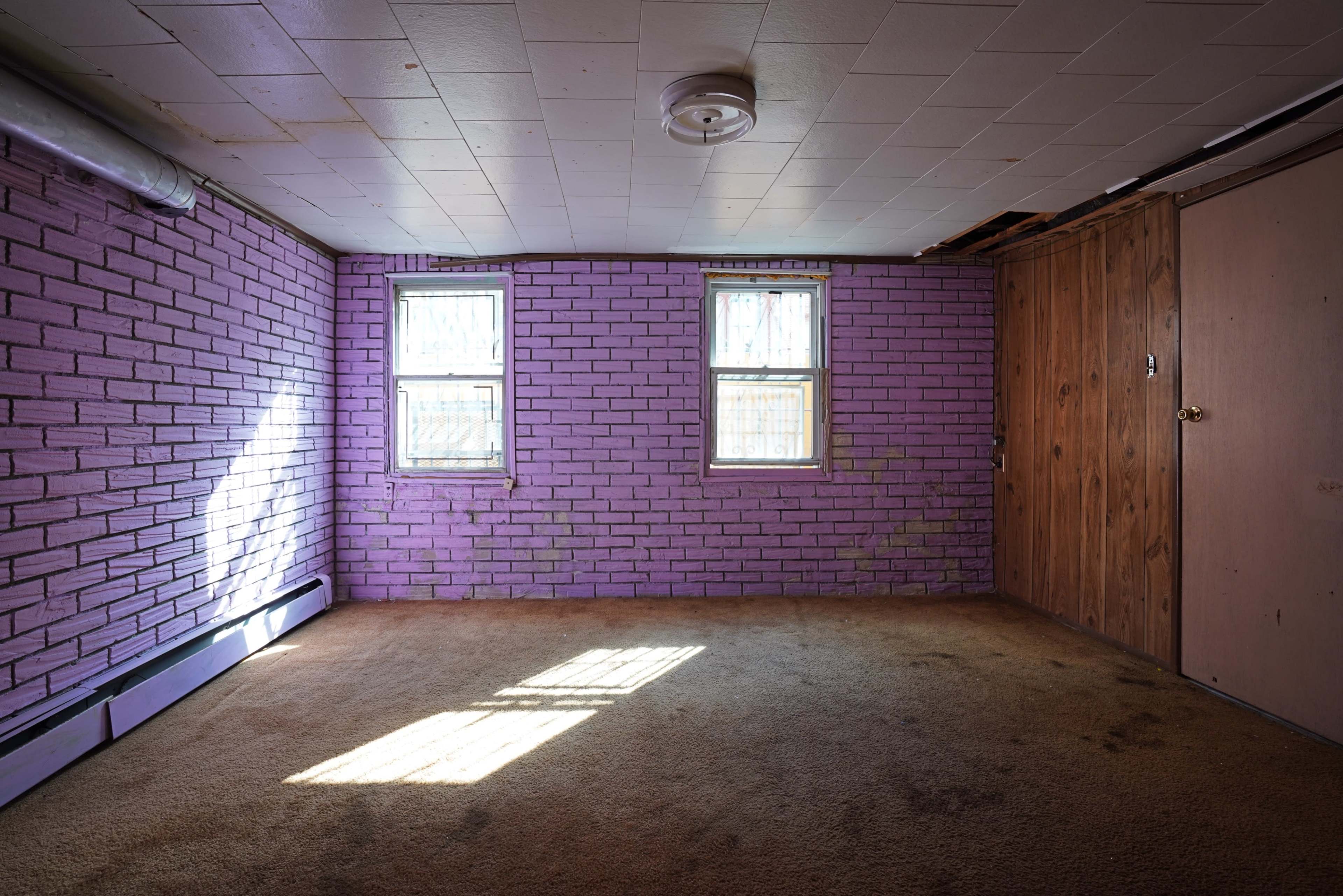 A sparsely furnished room with purple brick walls, two windows letting in natural light, and a wooden door on one side.