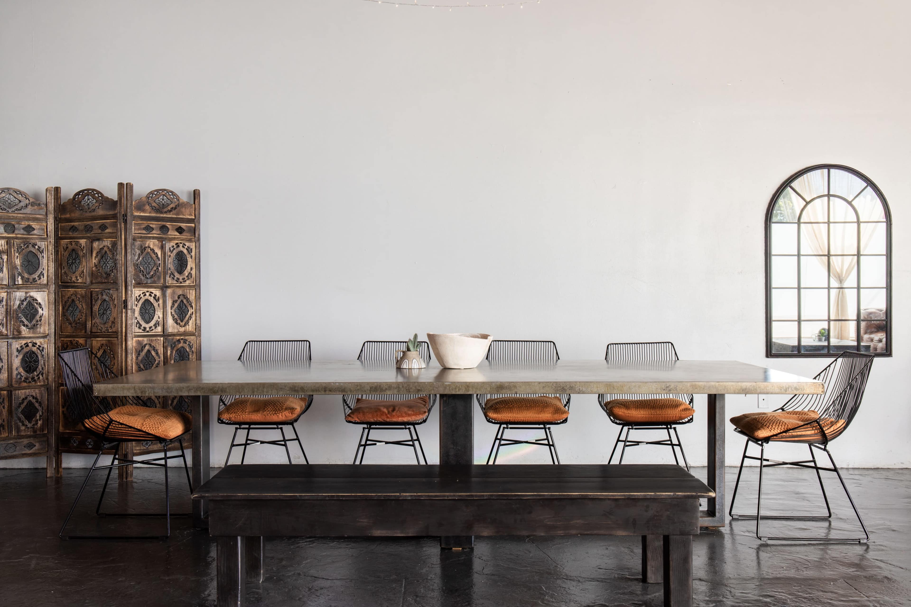 A large, rectangular dining table with metal chairs and a long bench is set against a plain wall, beside a decorative screen and a window.