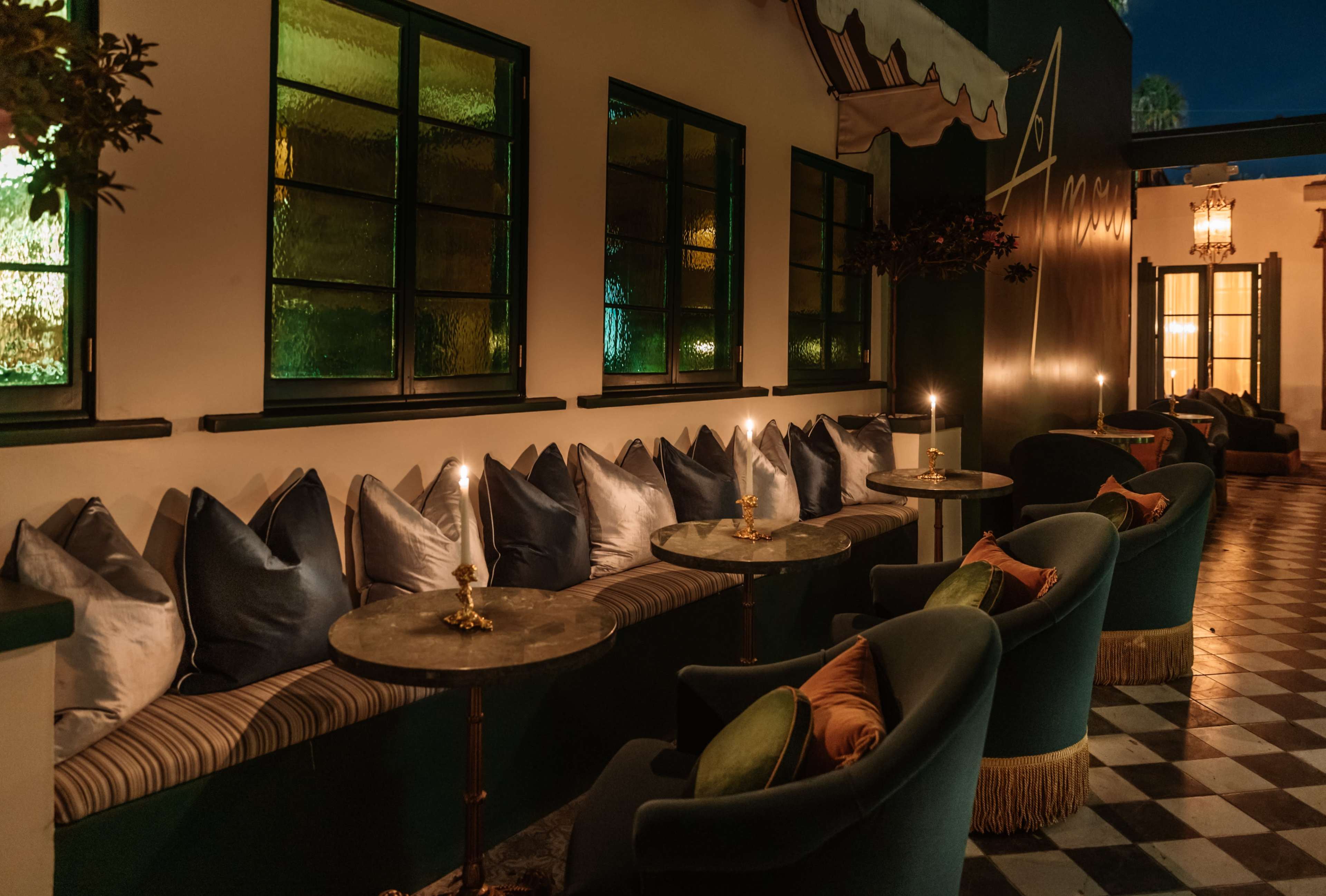 The image shows a cozy outdoor seating area featuring plush chairs with decorative pillows, small tables, and candles, against a backdrop of illuminated windows.