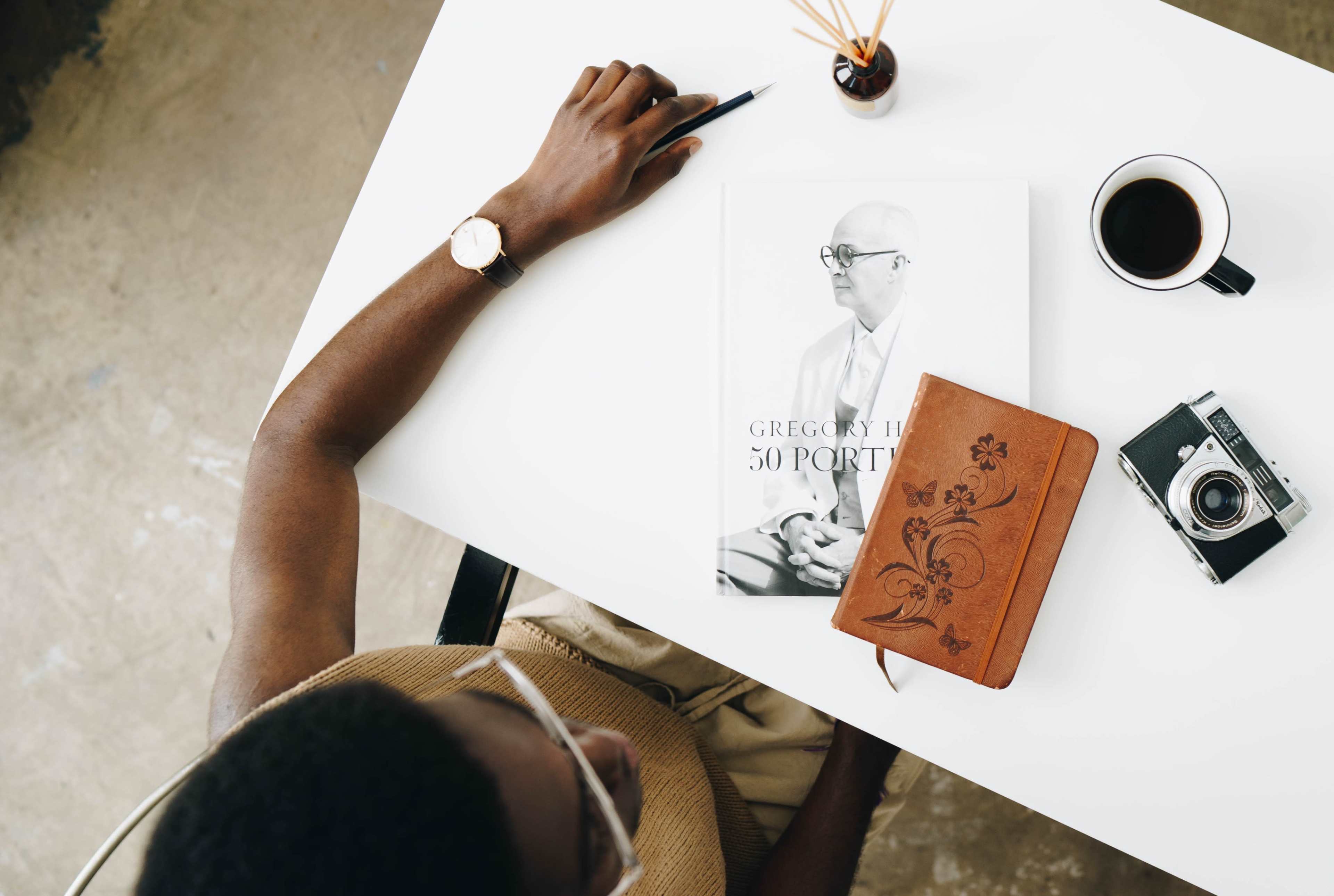 A person writes with a pen on a white table featuring a book titled "Gregory Halves 50 Portraits," a leather notebook, a camera, a cup of coffee, and a plant.