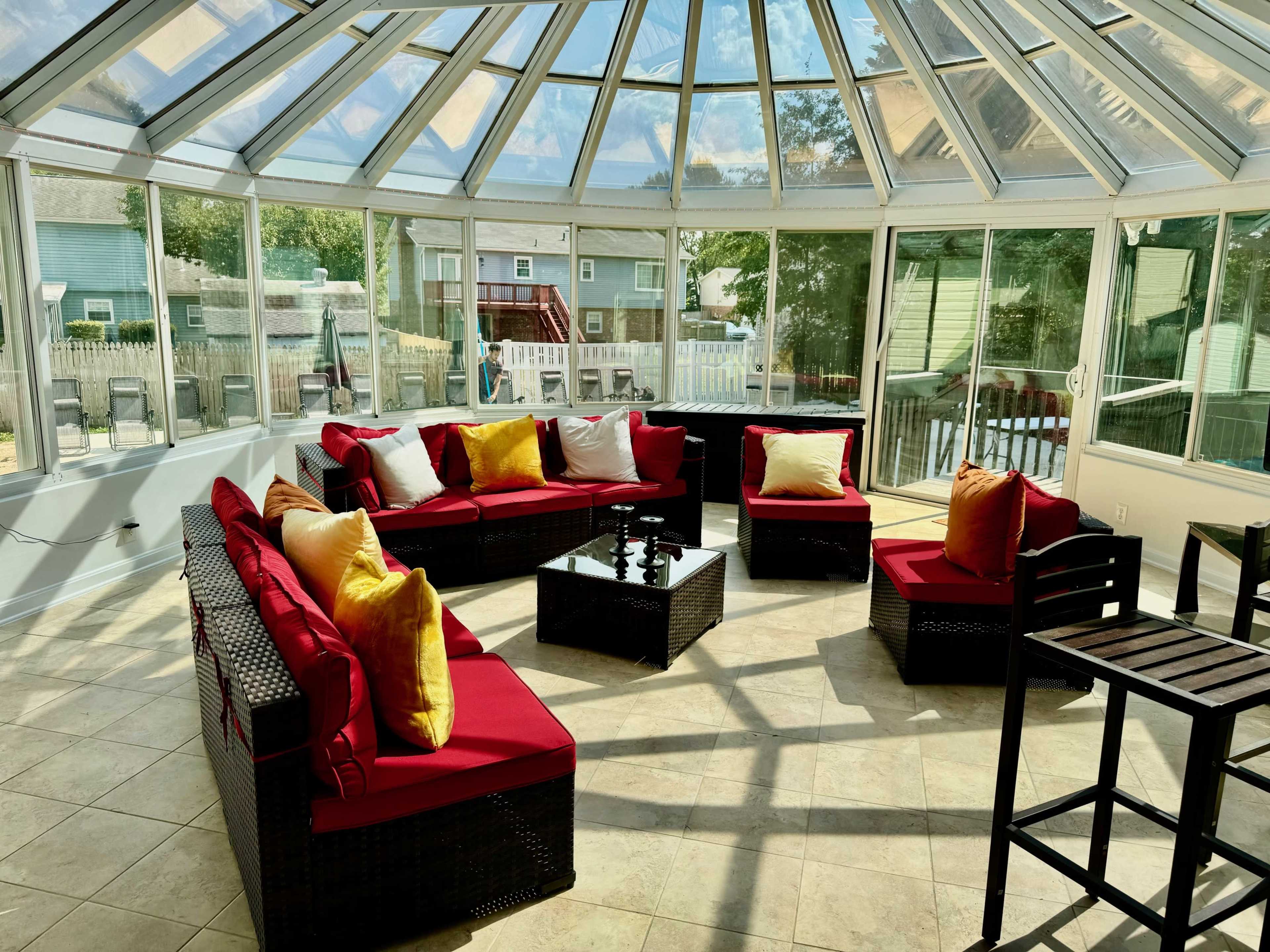 A bright sunroom with a circular arrangement of wicker seating, red cushions, and a glass table, surrounded by large windows that let in natural light.